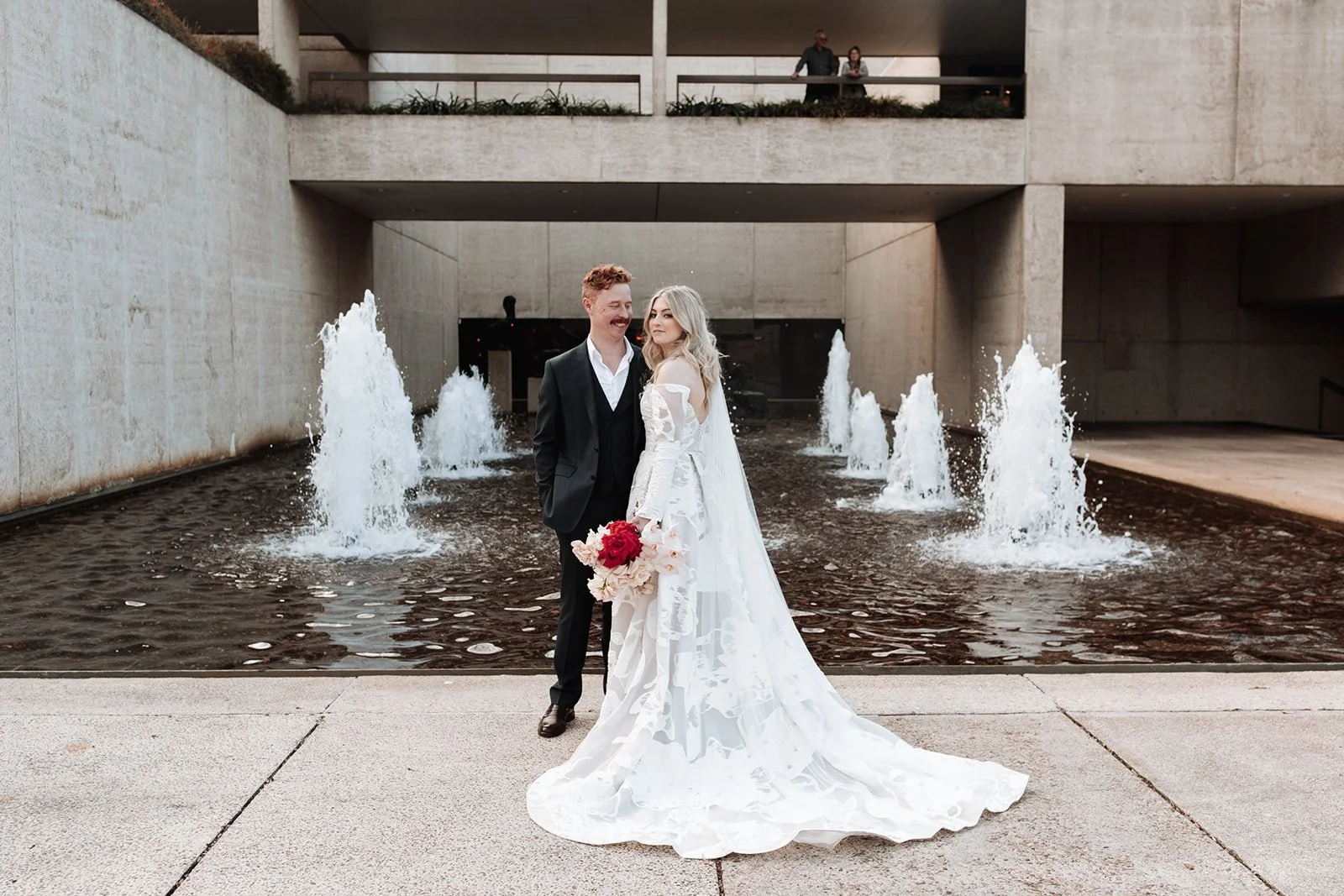 A newlywed couple stands in front of a water fountain at an urban location. The bride is wearing a white wedding gown and holding a bouquet of red and white flowers; the groom is in a black suit at a wedding at the queensland state library