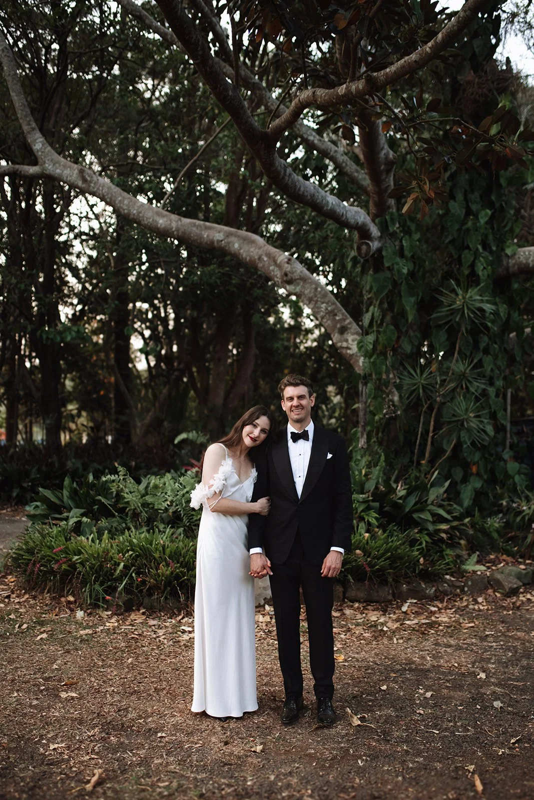 A bride and groom stand together outdoors amidst lush green trees and plants, holding hands, dressed in wedding attire, with the bride in a white gown and the groom in a black tuxedo, smiling after their wedding at Austinvilla Estate.