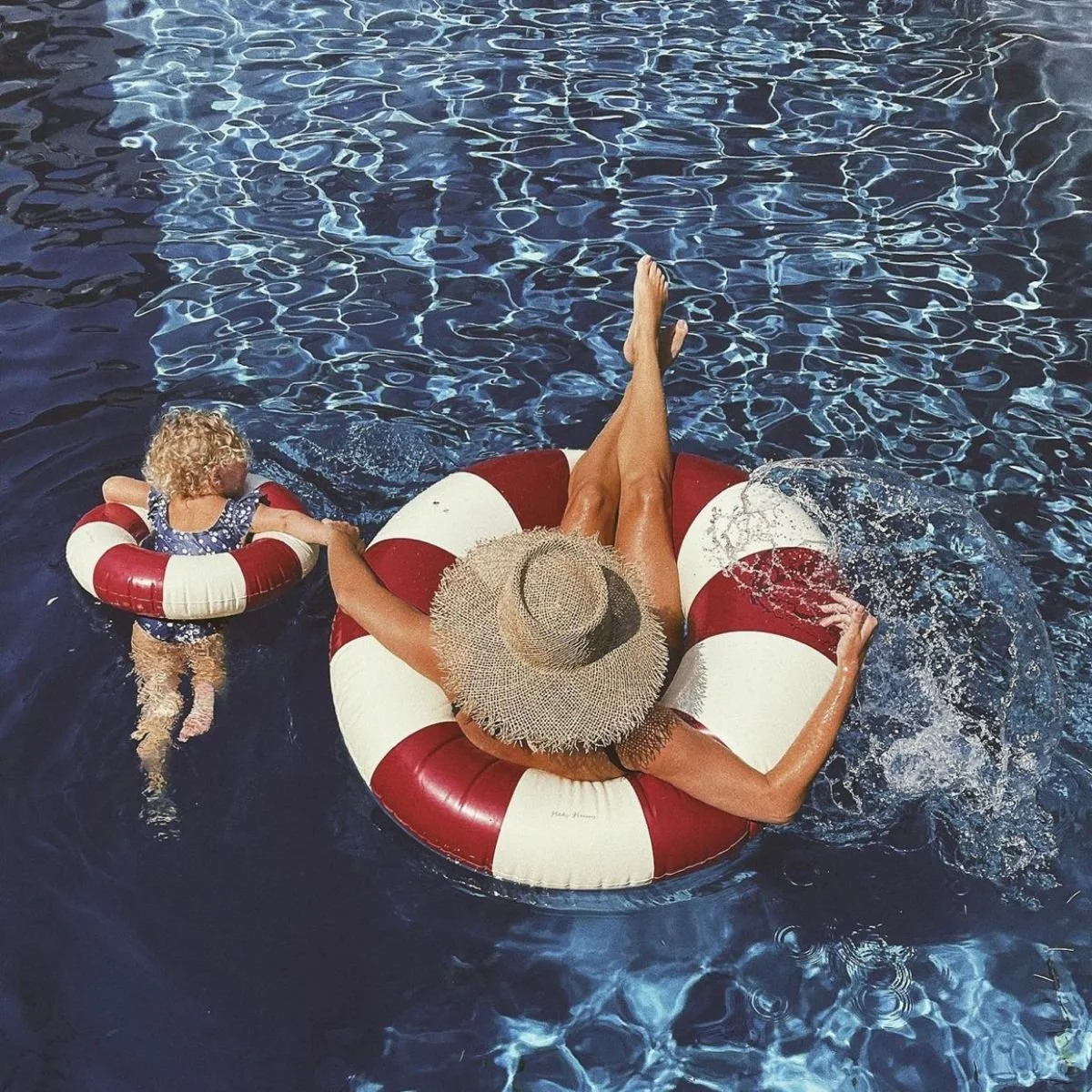 A woman in a straw hat relaxing on a red and white inflatable float with her legs crossed, holding hands with a young girl in a life jacket, both floating in a swimming pool with clear water.