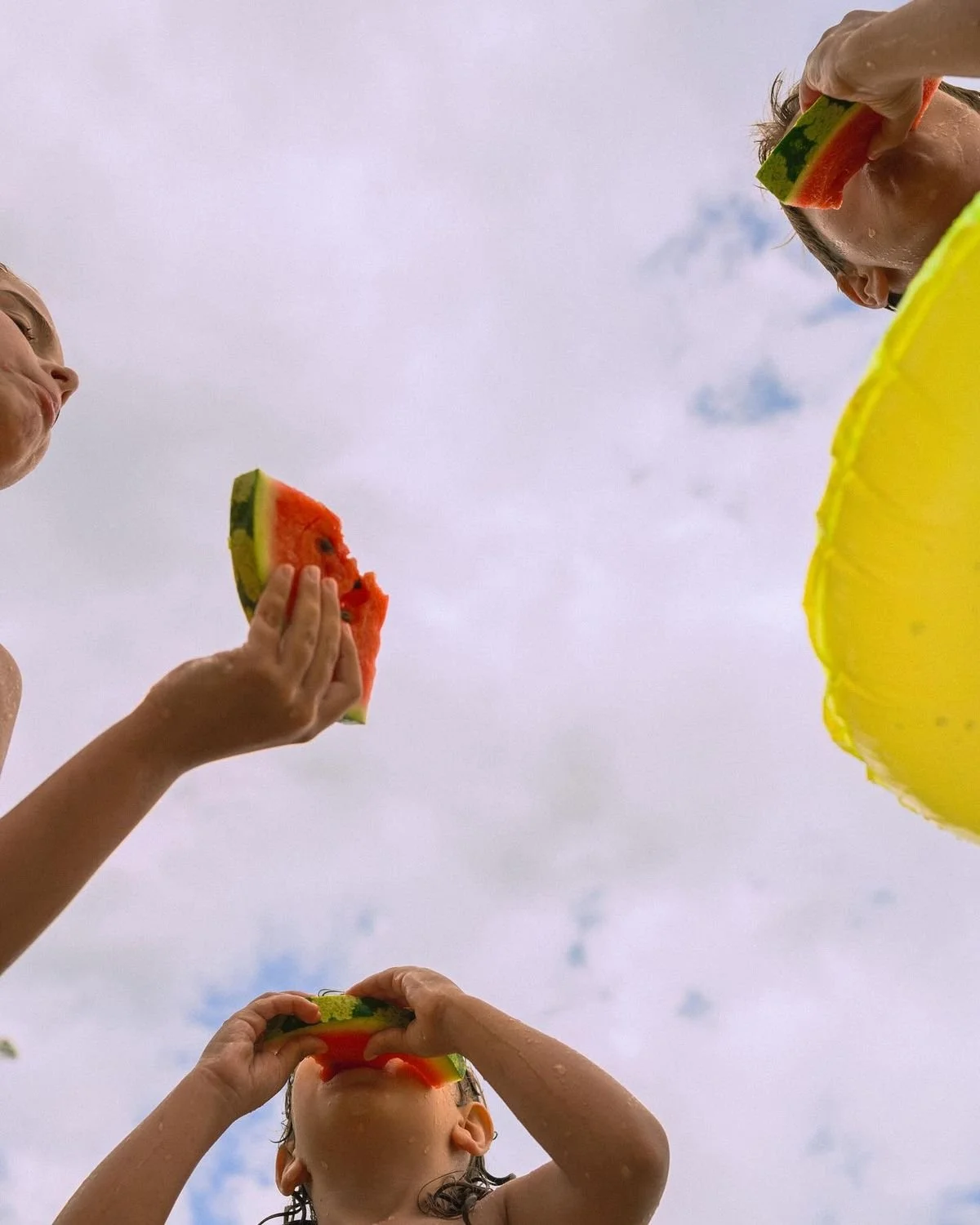 Three children are eating slices of watermelon outdoors on a cloudy day, viewed from below.