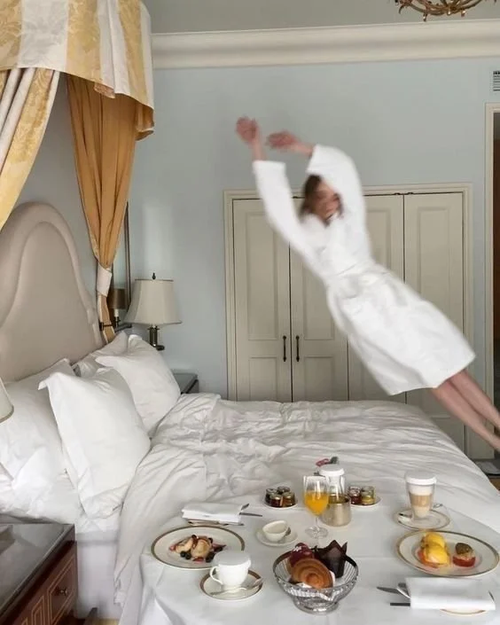 A woman in a white bathrobe is jumping on a hotel bed in a hotel room. The bed has white bedding and pillows, and there are breakfast items including coffee, juice, pastries, and fruit on the bed.