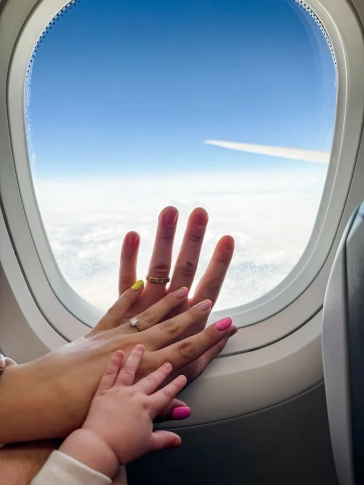 Two adult hands and one child's hand touching a plane window showing clouds and sky outside with a streak of airplane contrail.