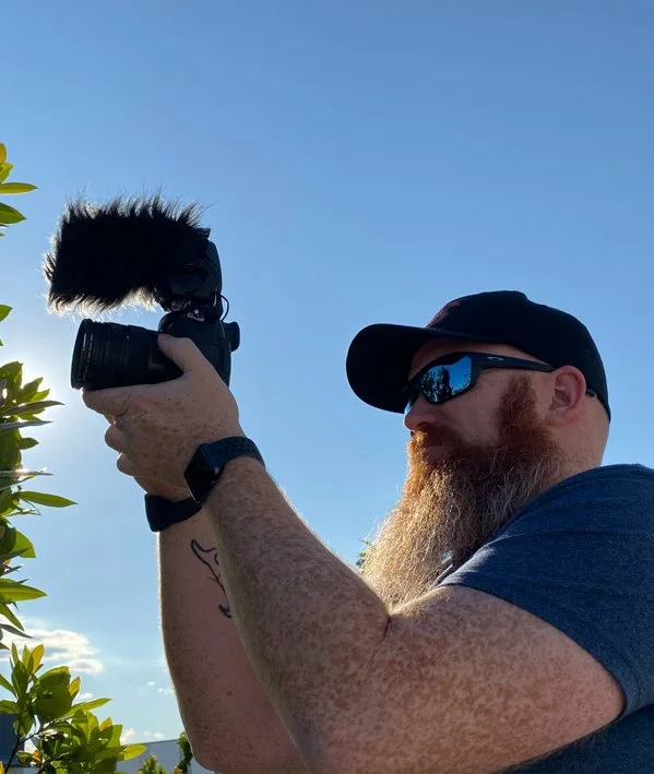 Man with a beard and sunglasses holding a camera with a windshield and fuzzy microphone, outdoors with clear blue sky and green foliage.