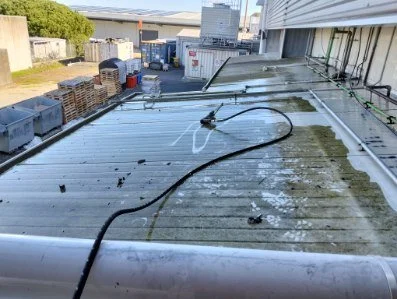 Cleaning a flat metal rooftop with a pressure washer, with buildings and storage containers in the background.
