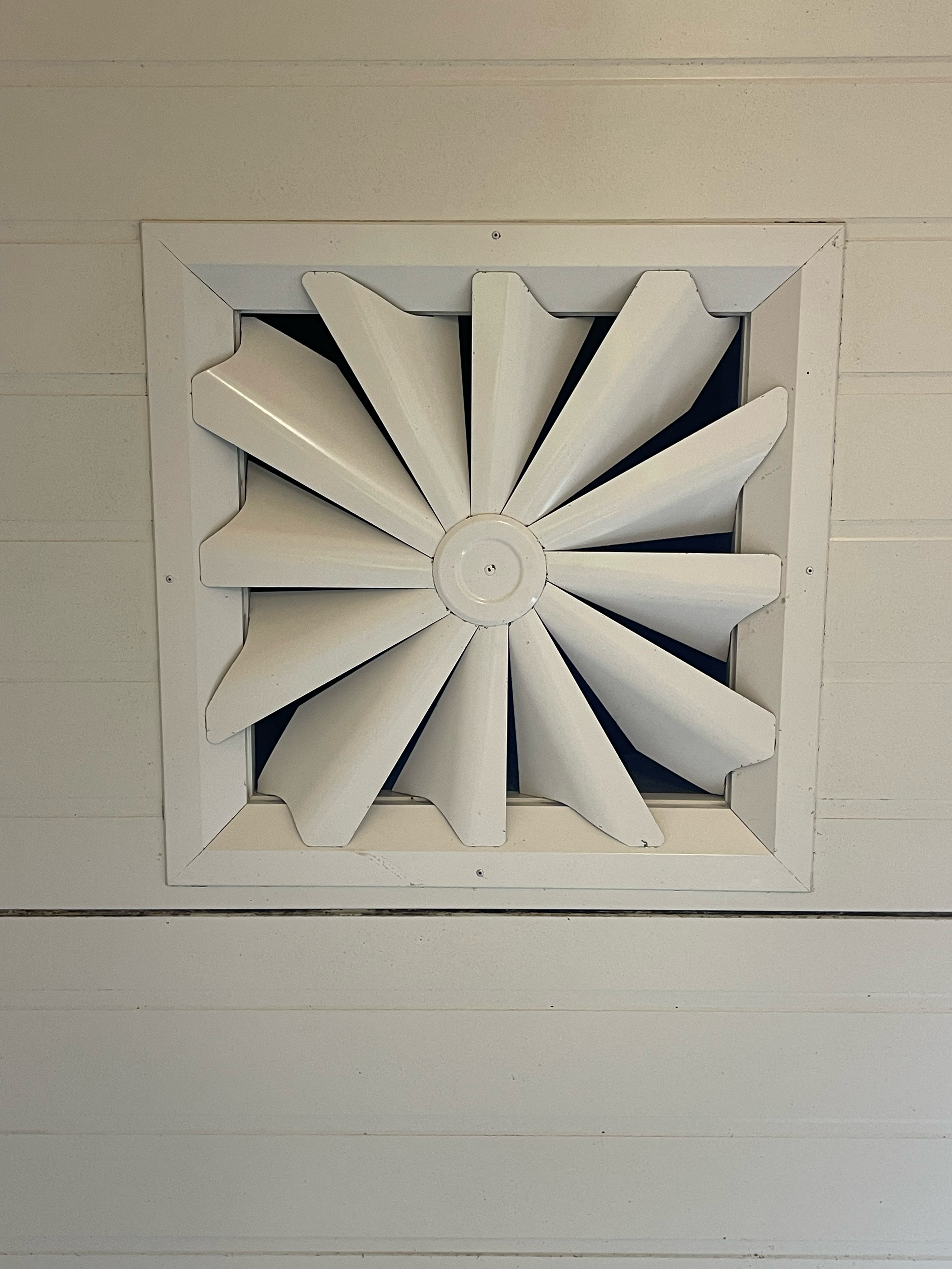 A white square vent cover with a decorative design resembling a pinwheel or spiral, mounted on a white wooden wall.