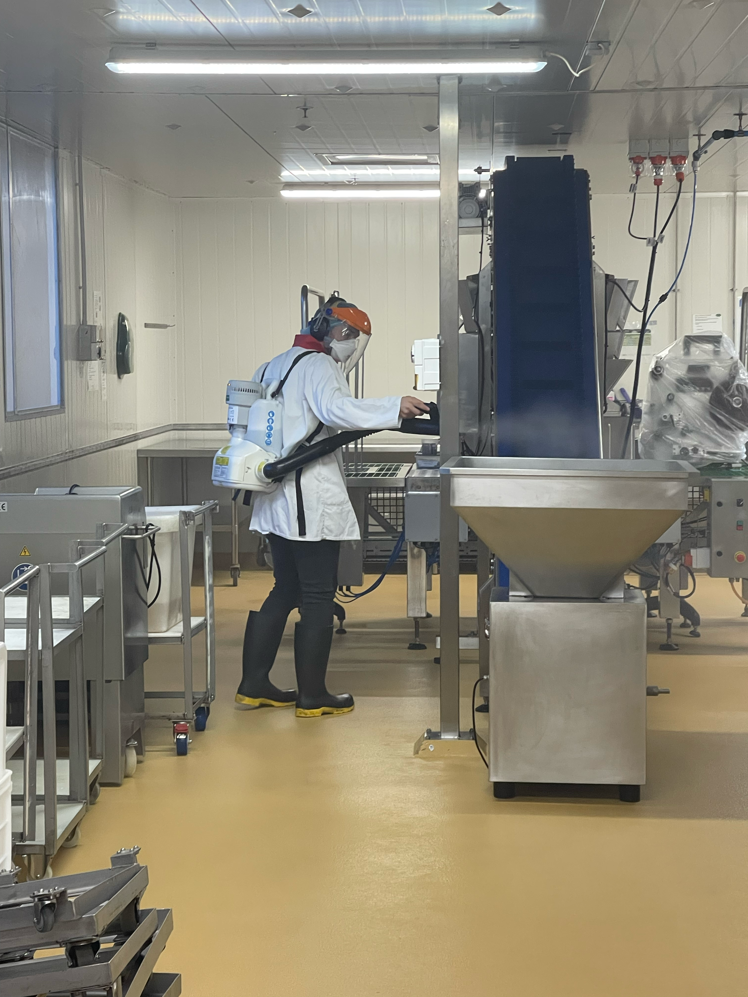 A worker in protective gear, including a face shield, mask, white gown, gloves, and slippers, operating machinery in a clean industrial or laboratory setting.