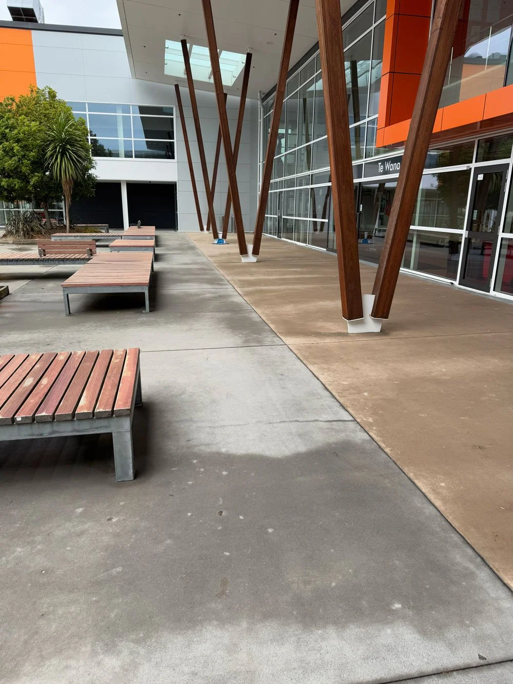 Modern outdoor building entrance with wooden support beams, large glass windows, and wooden benches on concrete and tiled pavement.