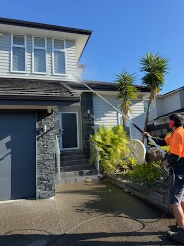 A person spraying water on a house exterior with a hose, standing on a wet driveway in front of a modern house with a gray garage door and white siding, and a small garden with palm trees.