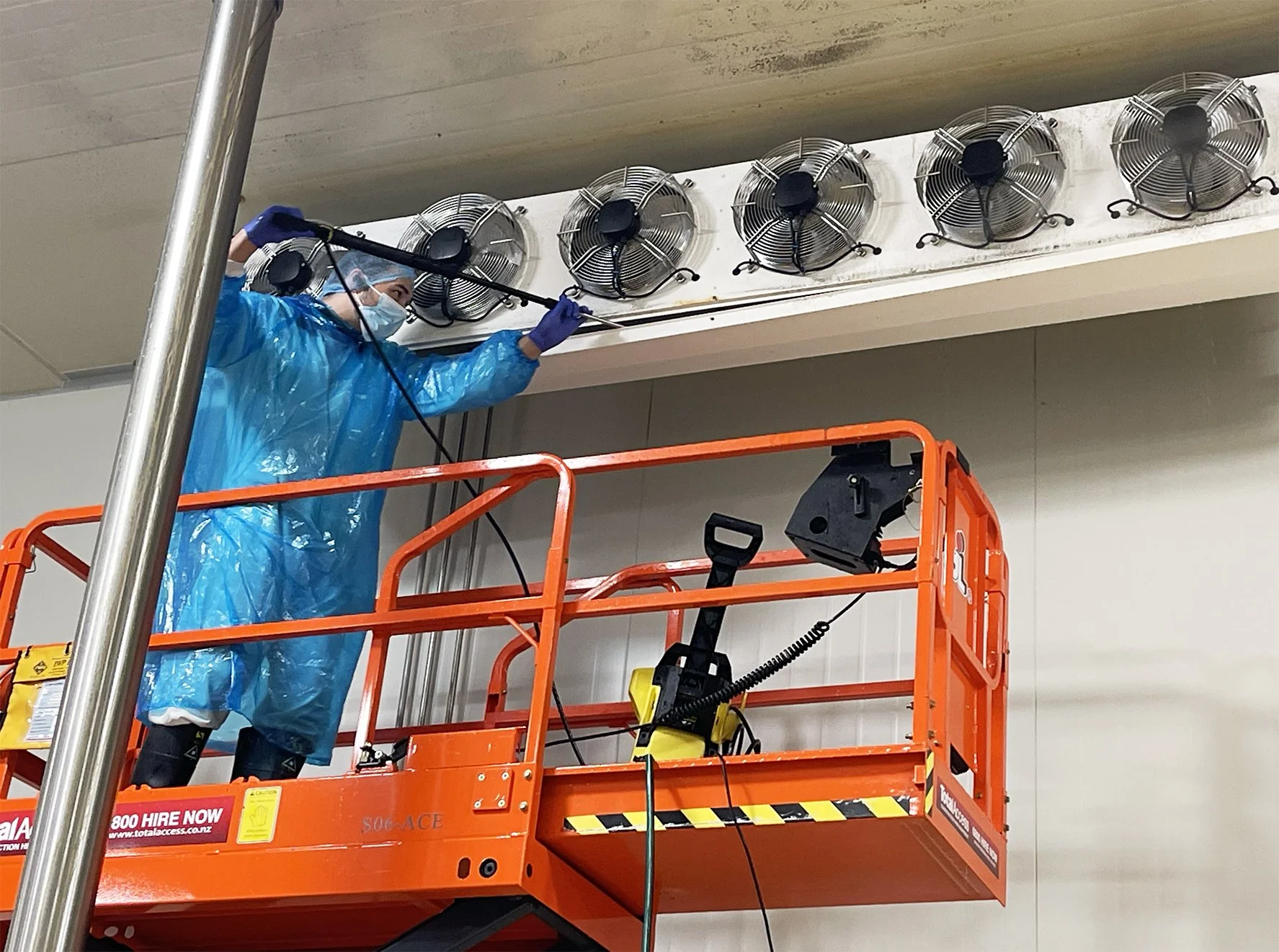 Worker in blue protective gear, face mask, and gloves on an orange lift, cleaning or inspecting industrial ceiling fans in a large indoor space.