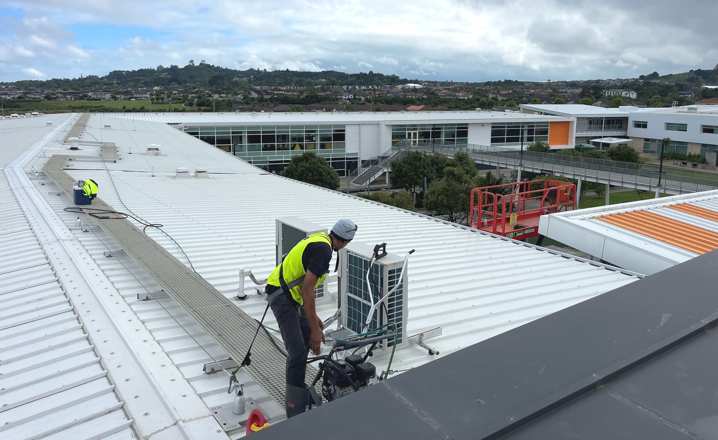 A worker on a white metal roof inspecting or maintaining solar panels with tools and equipment. The background shows a modern building with large glass windows and a walkway, with a residential neighborhood and green hills in the distance under cloudy skies.