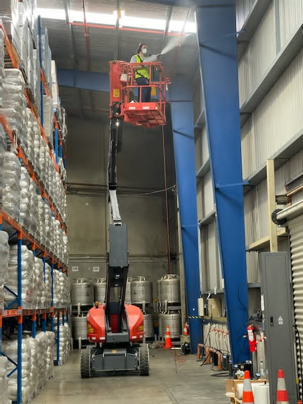 Worker in safety gear on a cherry picker's platform cleaning the warehouse ceiling.