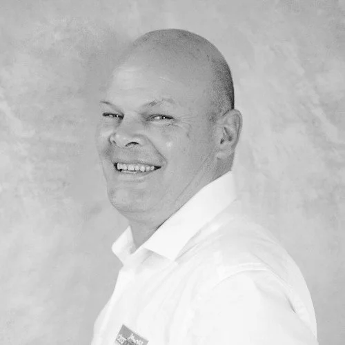 Black-and-white photo of a smiling man with a bald head, wearing a white collared shirt.