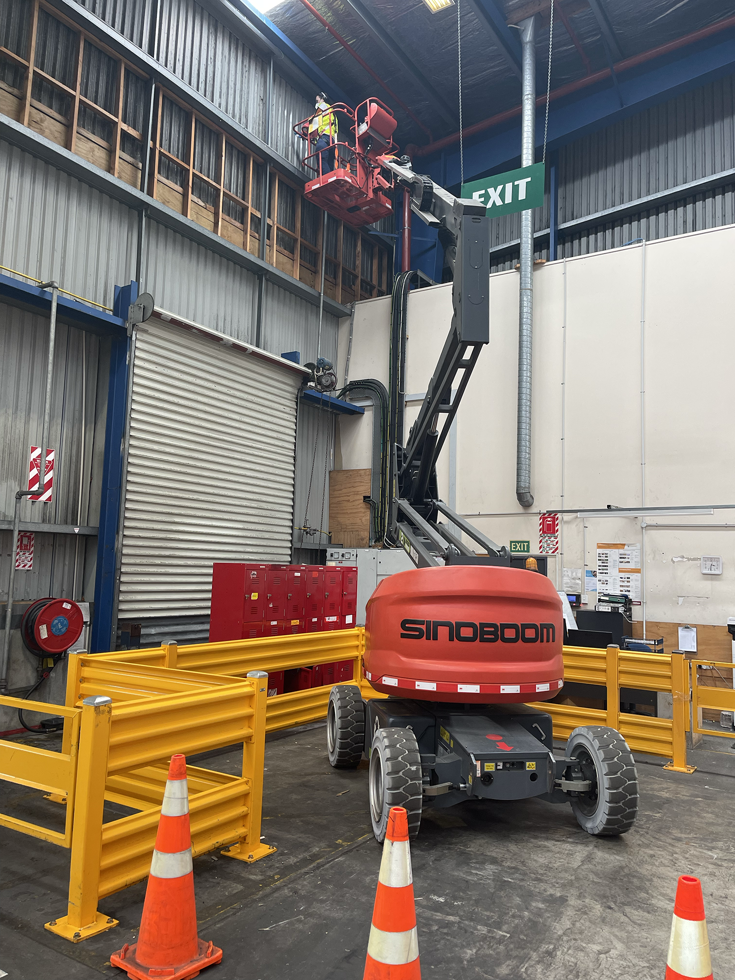 Indoor construction site with a man operating a red elevated work platform, surrounded by safety barriers and traffic cones, with exit signs and construction materials in the background.