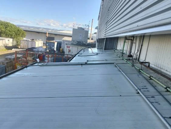 View of a building rooftop with cables, pipes, and metal siding, overlooking an industrial area with containers and structures under a partly cloudy sky.