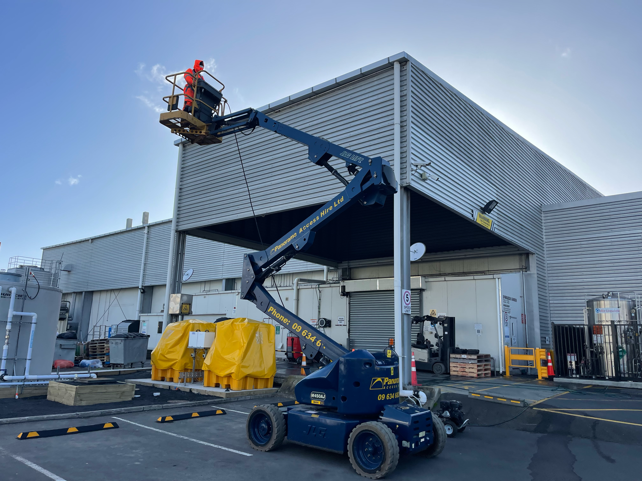 A worker in a red safety suit is on a blue cherry picker lifting towards the corner of a commercial building with metal siding. The scene takes place in a parking lot with various supplies, warning signs, and equipment around.