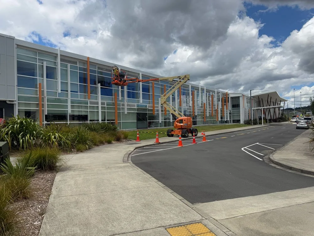 A construction lift working on a modern building with glass windows, surrounded by orange cones and caution tape, on a cloudy day.