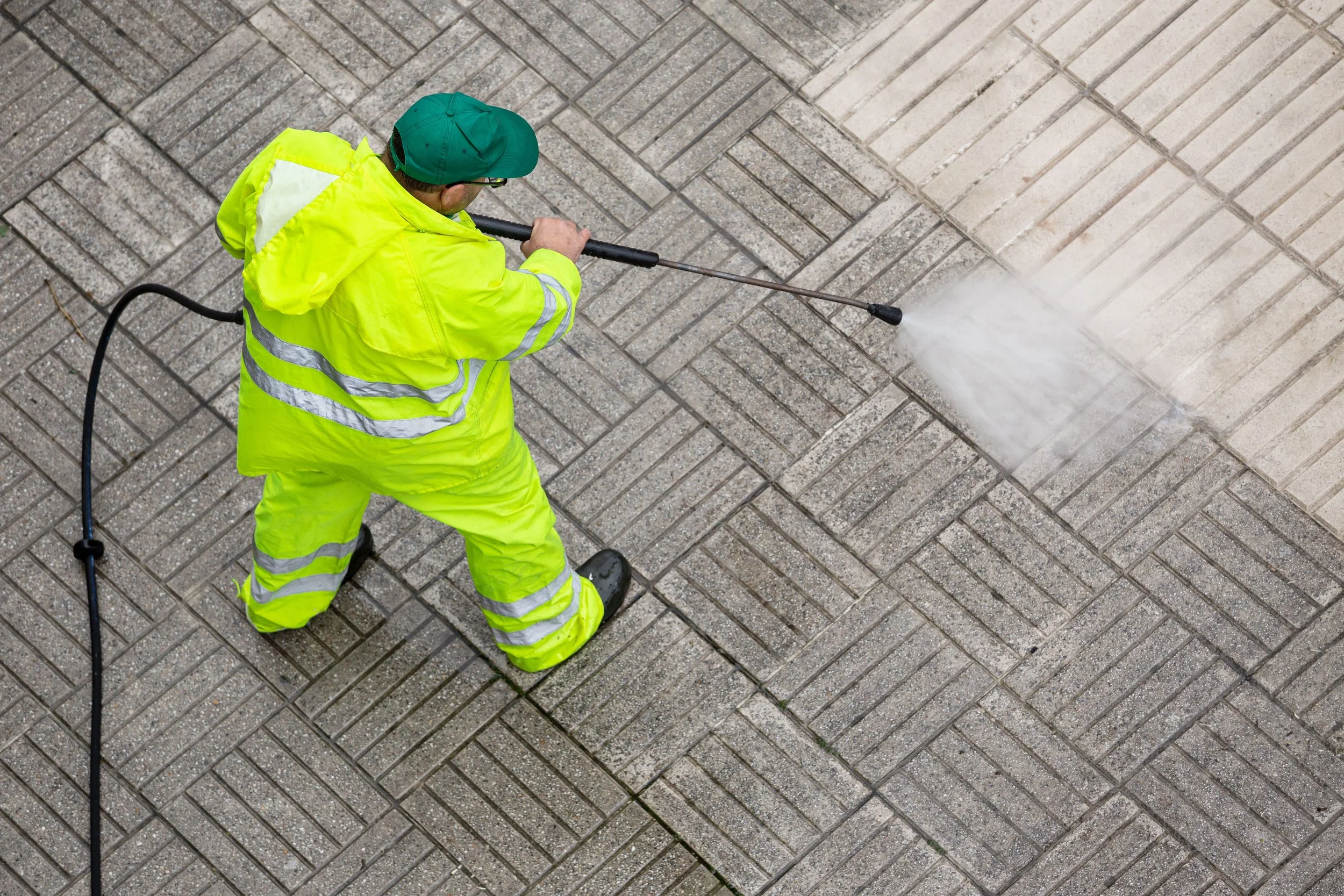 A person in a high-visibility yellow jacket and pants, wearing a green cap and glasses, cleaning a brick sidewalk with a pressure washer.