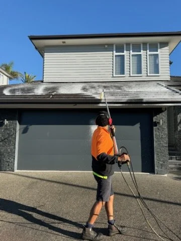 A person in an orange shirt and black shorts cleaning the roof of a two-story house with a power washer, standing on the driveway.