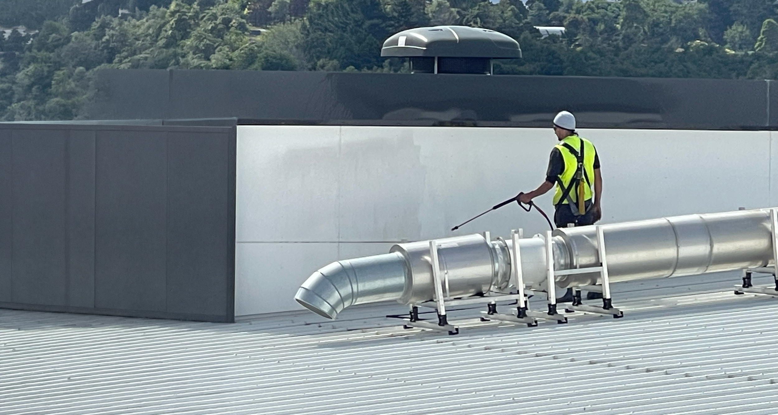 A worker in a neon yellow safety vest, white hard hat, and sunglasses, standing on a rooftop with ventilation equipment, using a spray for cleaning or maintenance, with a backdrop of trees and a partly cloudy sky.