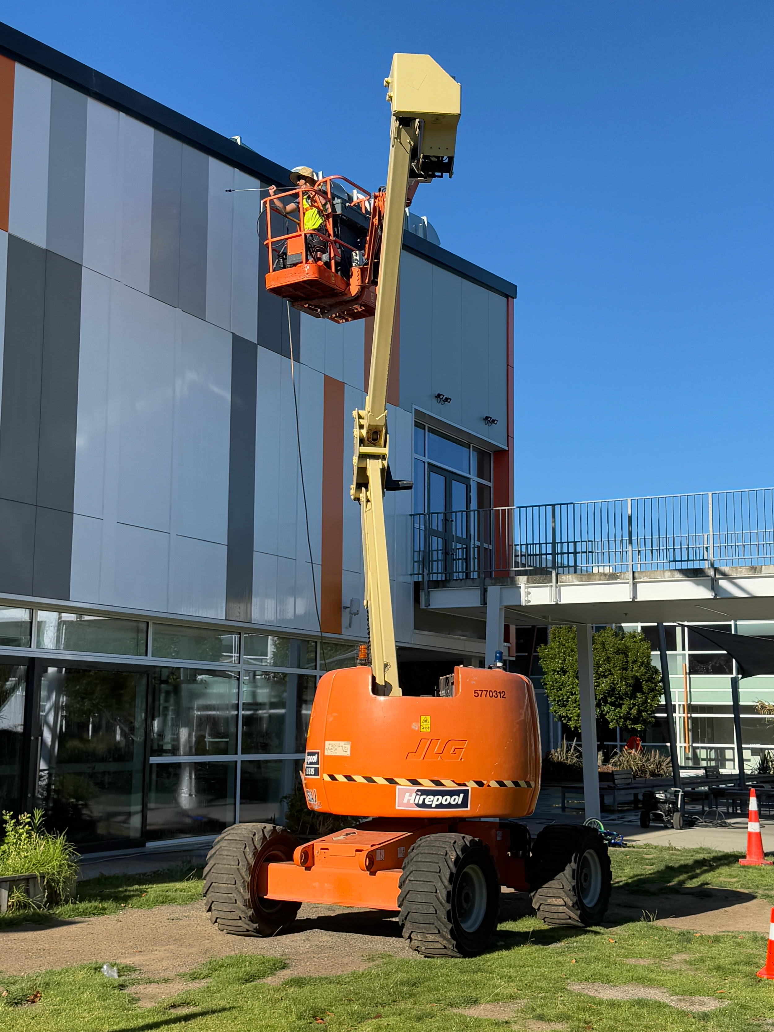 Construction worker on an orange cherry picker painting or repairing the exterior of a modern building with glass windows and gray, white, and orange panels.