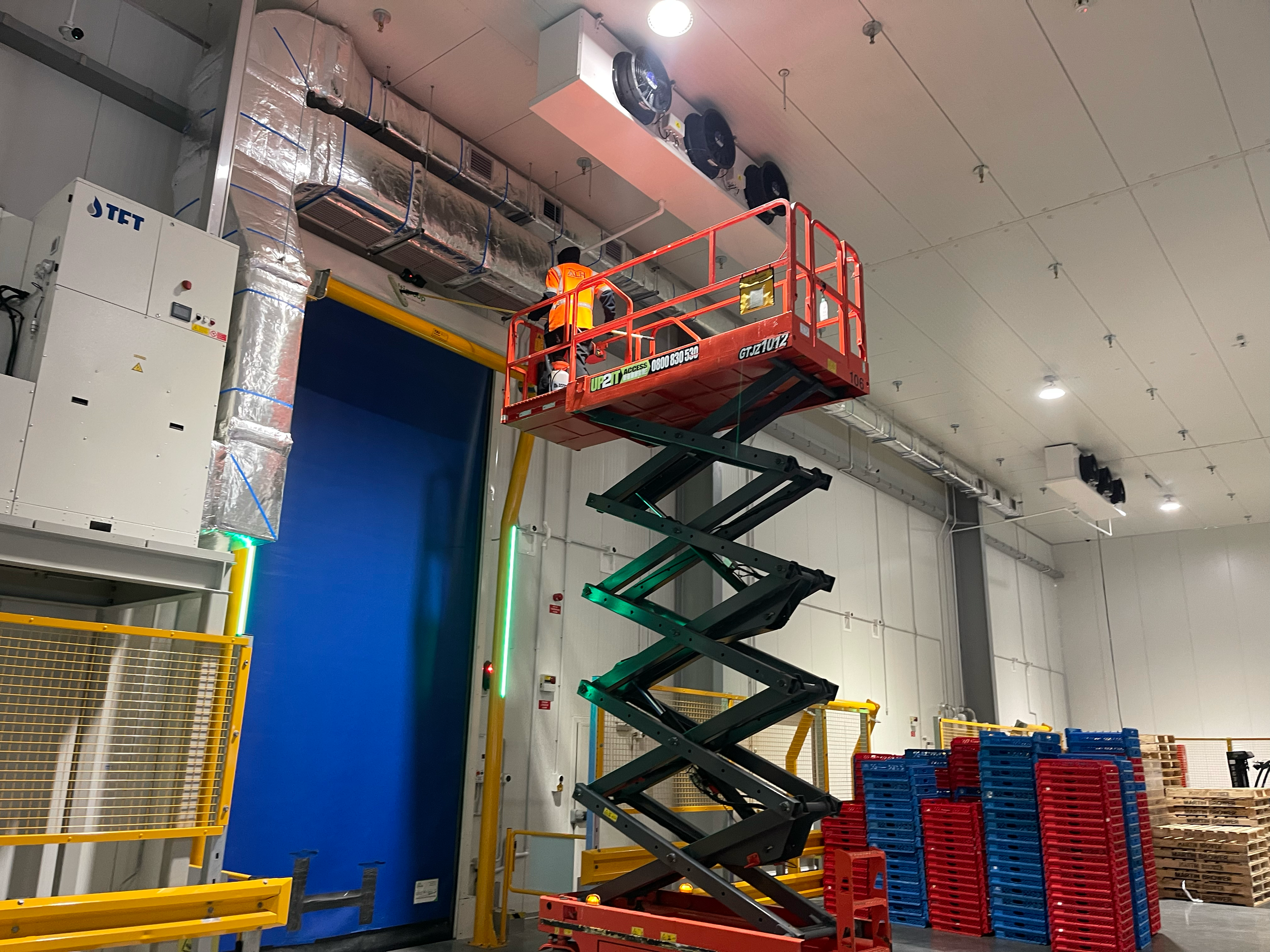 A worker wearing an orange safety vest and helmet is on a scissor lift working on HVAC ductwork in an industrial or warehouse setting. The ceiling has multiple vents and air conditioning units, with various striped insulation pipes and cables. There are stacks of red and blue plastic crates on the floor.