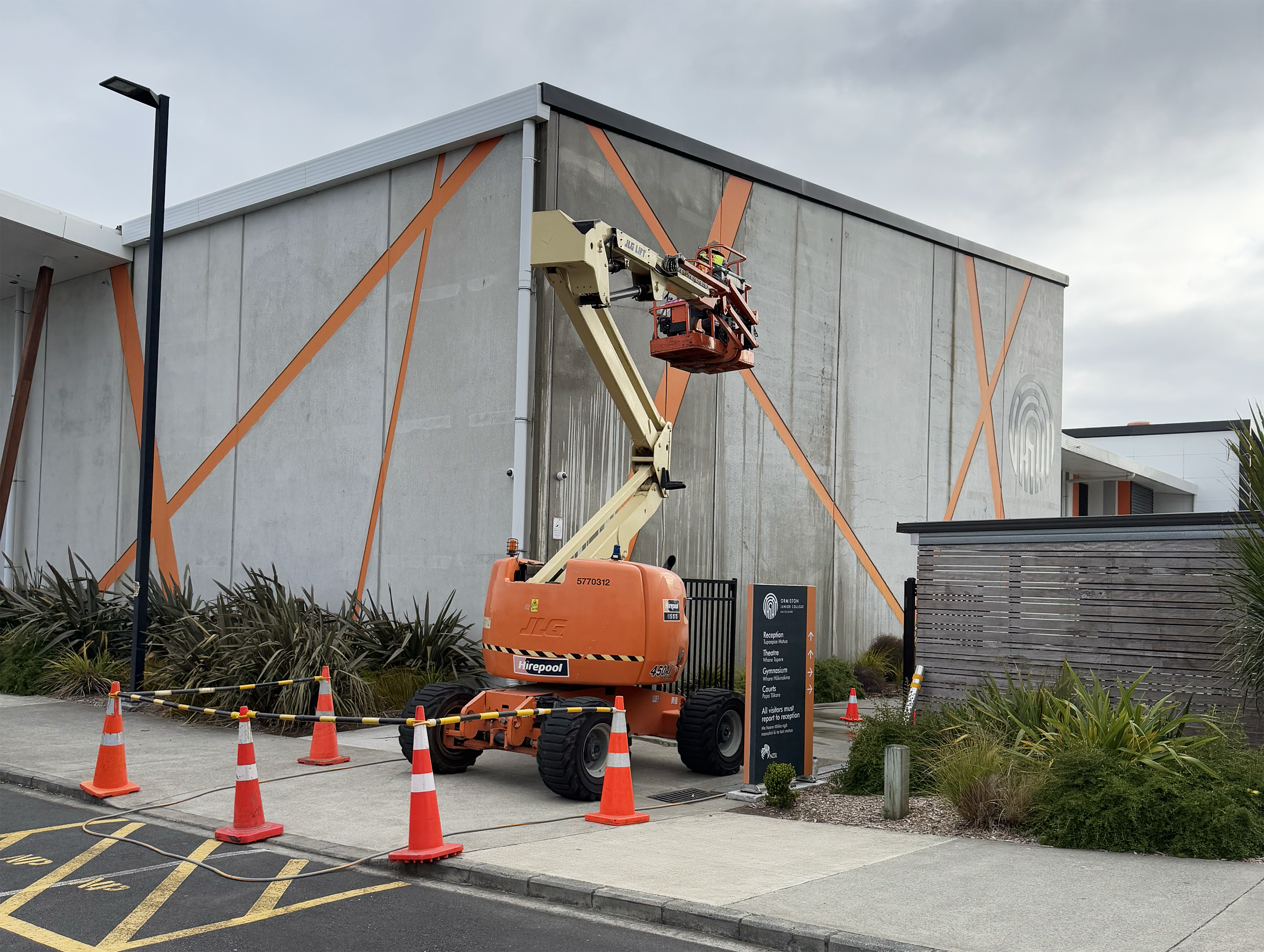 A construction lift with a worker on it next to a modern building with orange accents, surrounded by traffic cones and safety signs.