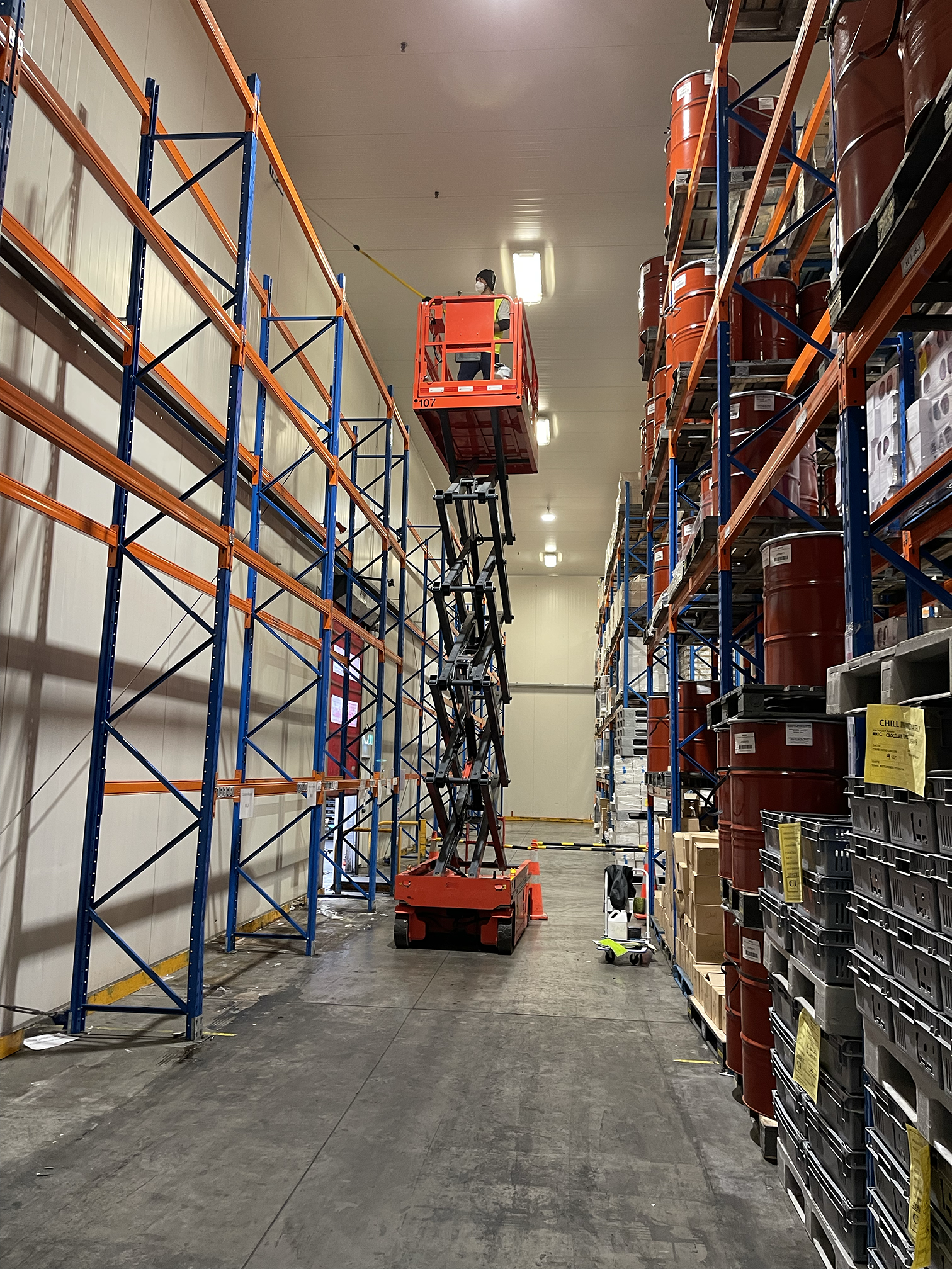 Warehouse aisle with blue and orange shelving units, and a worker on a cherry picker wearing a face mask.