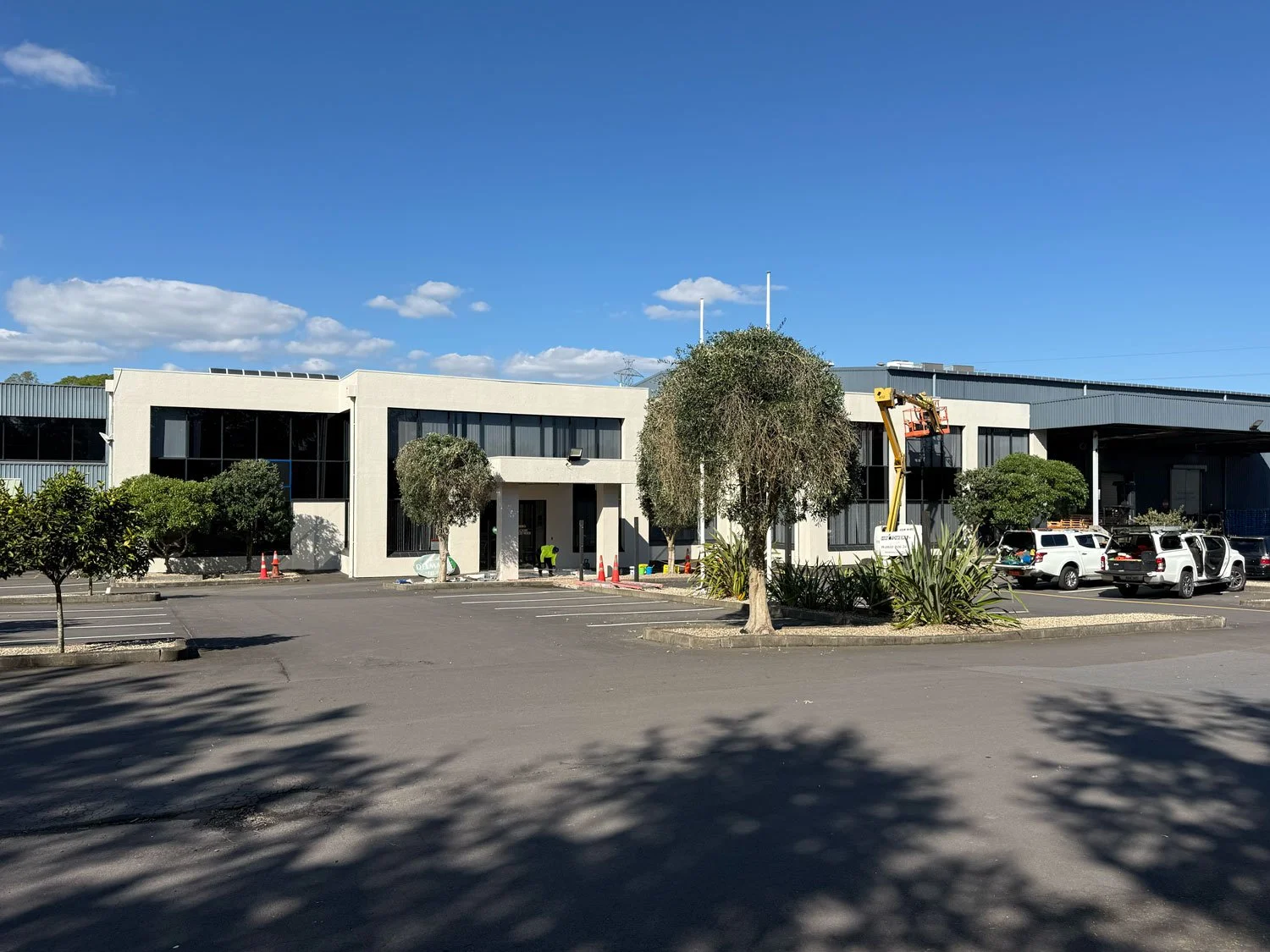 A modern commercial building with large windows, surrounded by small trees, parked cars, and construction workers preparing the entrance under a blue sky.