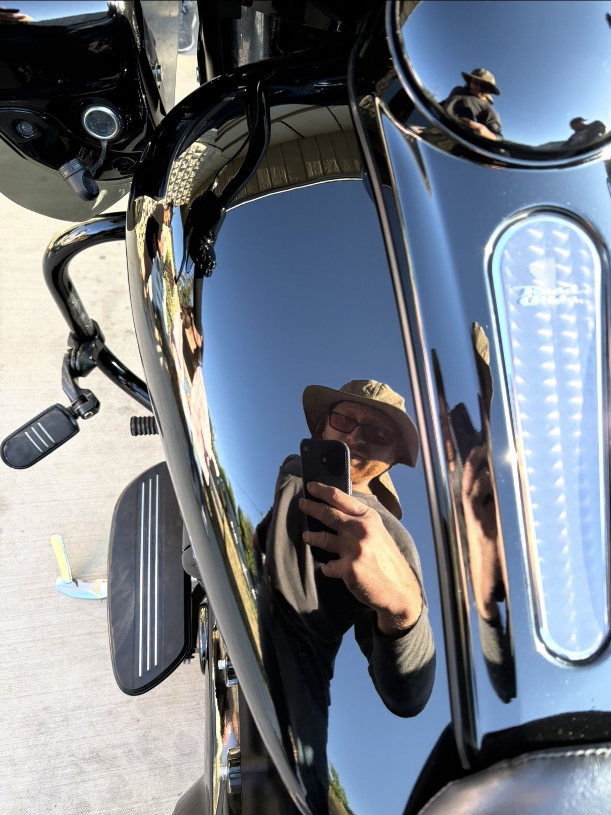 Reflection of a man wearing a hat and glasses taking a photo with his phone, seen on the shiny black surface of a motorcycle.