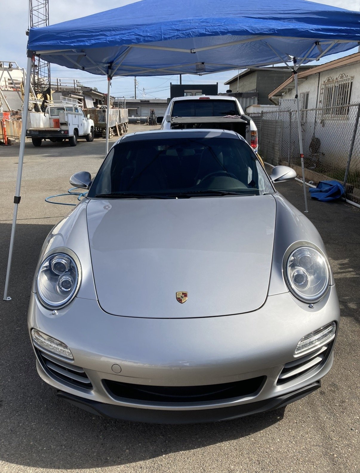 Silver Porsche sports car parked under a blue canopy tent on a gray asphalt lot, with a white truck and a fenced area in the background.