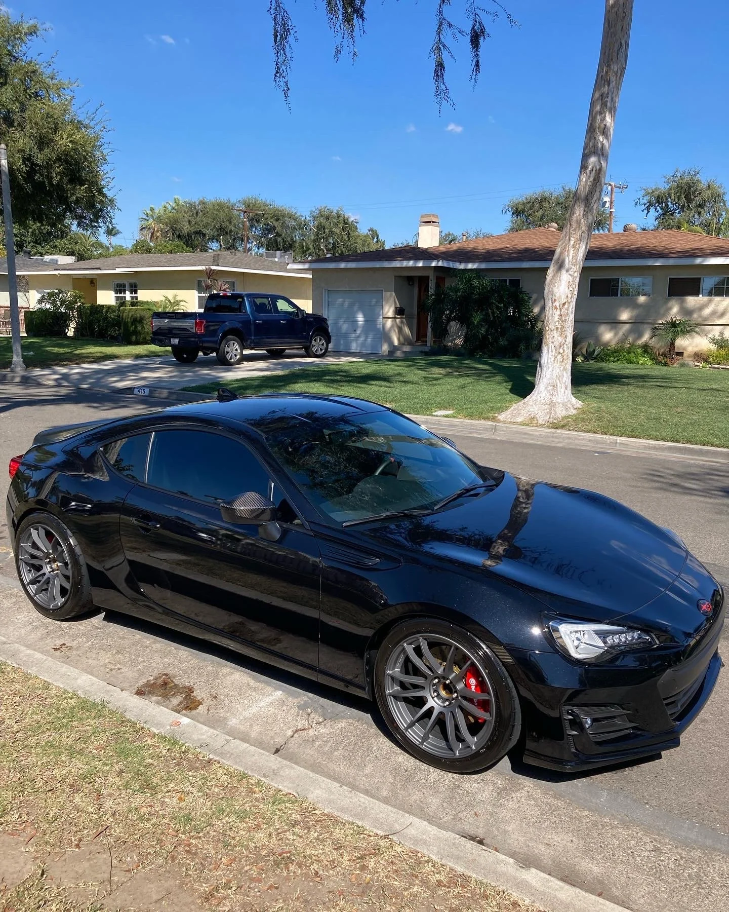 Black sports car parked on the side of the street in front of a grassy lawn and a house with a brown roof, trees, and a blue sky in the background.