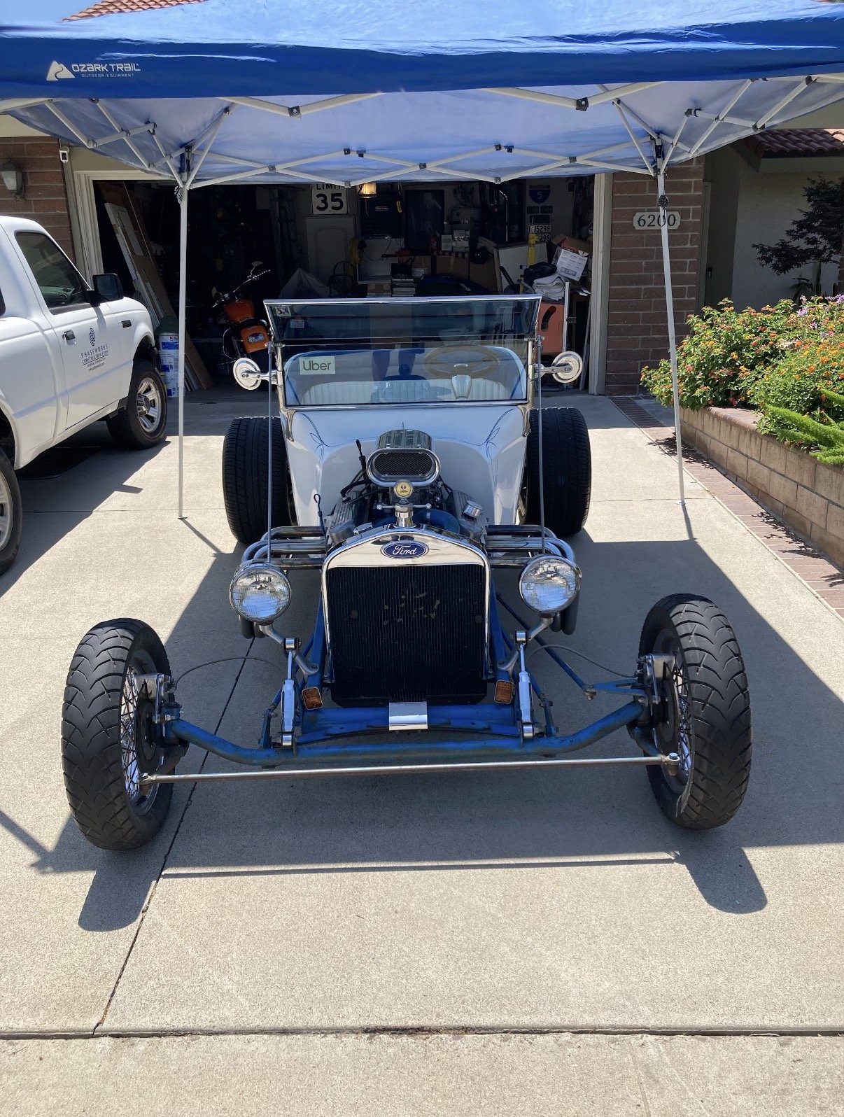 A vintage blue and white race car with a Ford logo, parked under a blue canopy in a driveway, with a garage and house in the background.