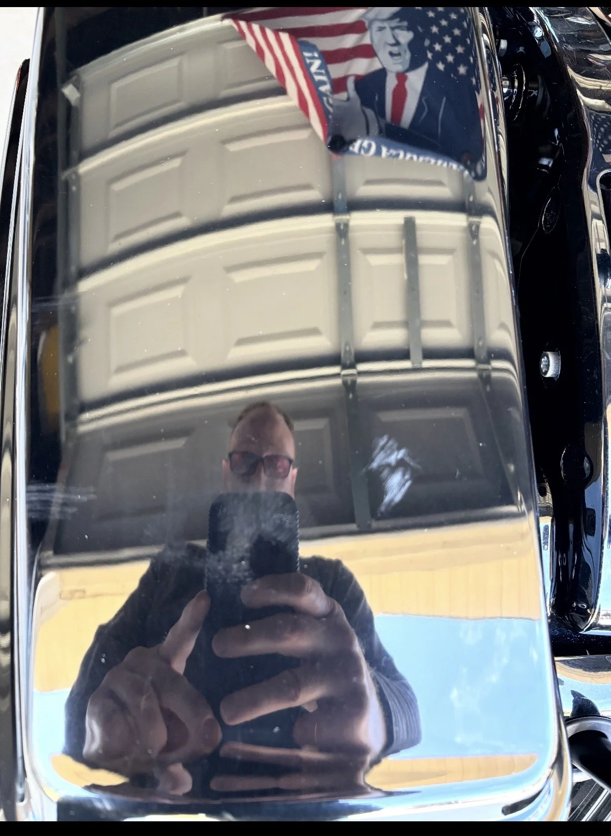 Reflection of a man taking a photo with a phone in a shiny, curved surface, possibly a vintage car's hood, during daytime.