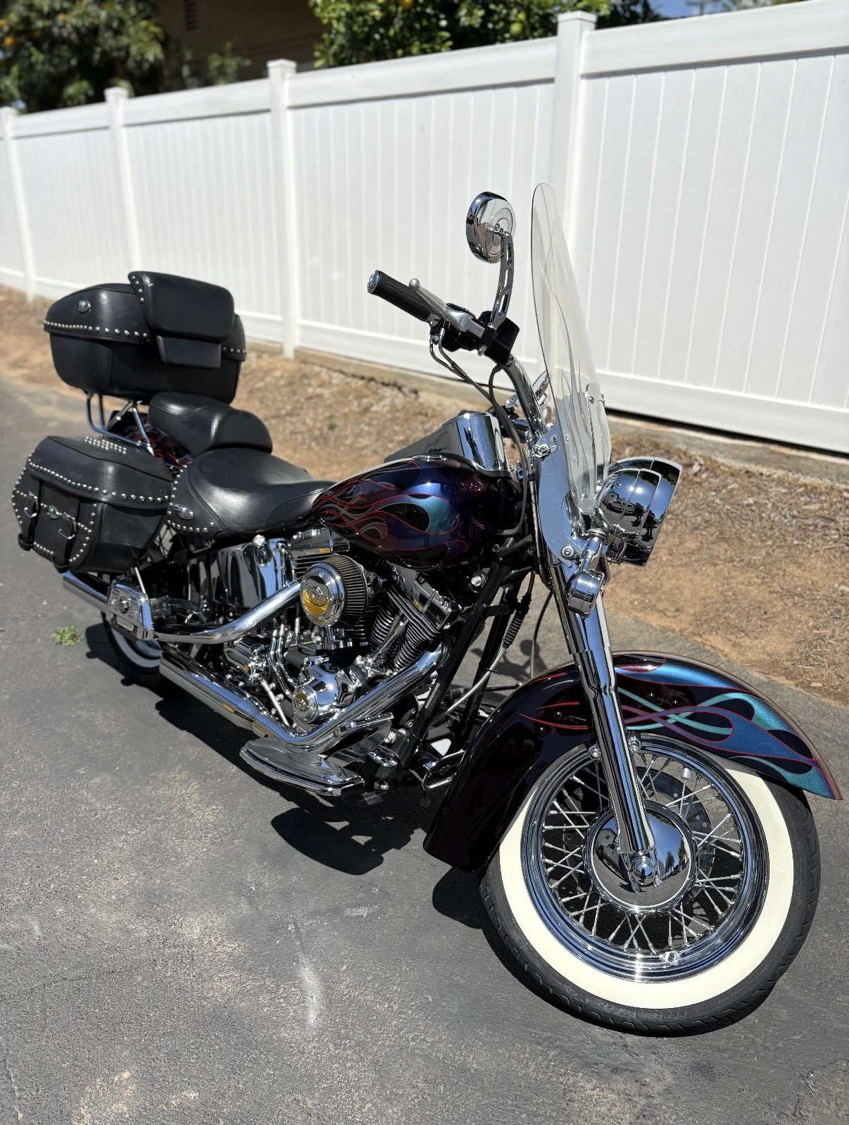 A black motorcycle with whitewall tires parked on a paved surface in front of a white fence. The motorcycle has chrome accents, a windshield, and black leather saddlebags with studs. The fuel tank has a flame design.