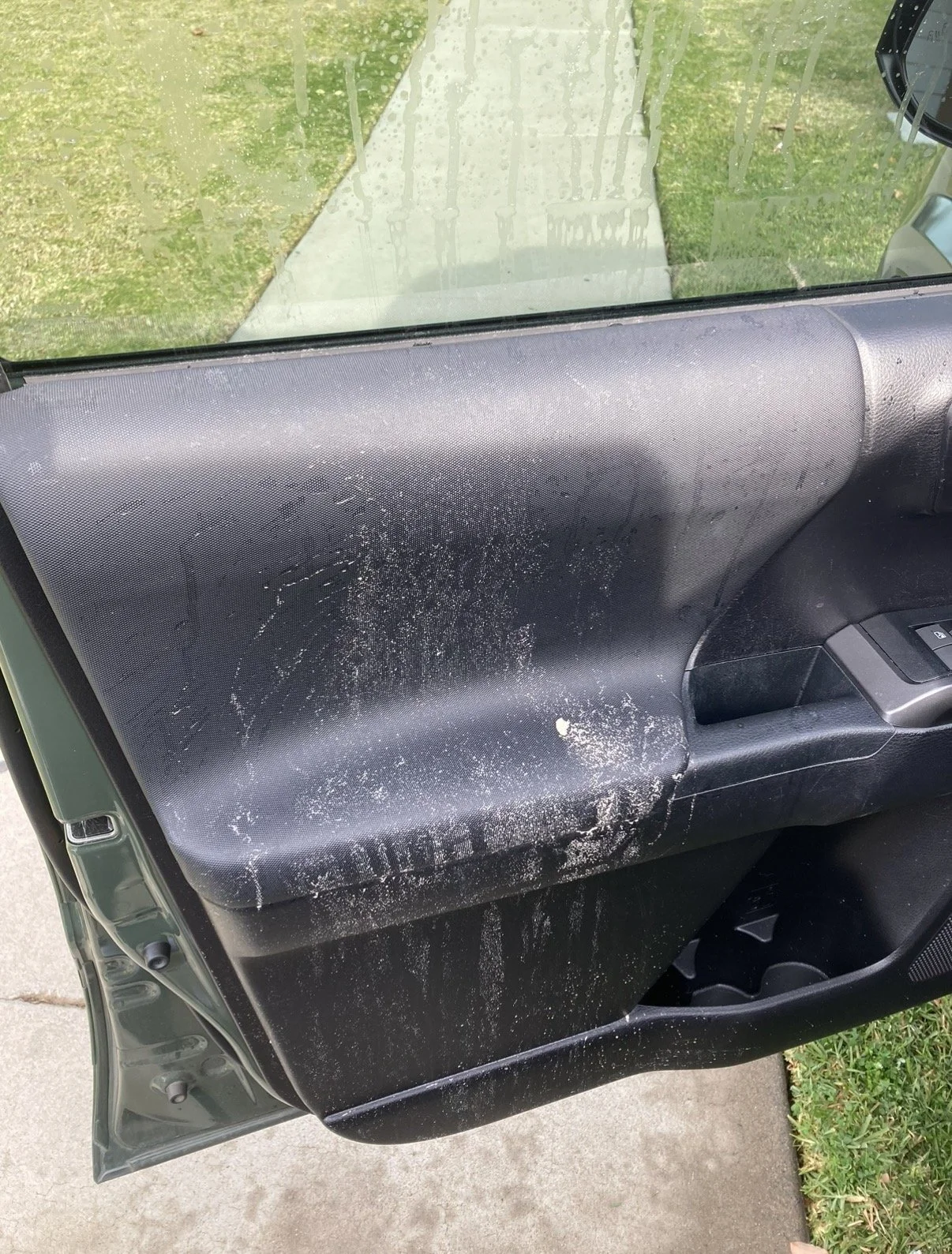 The inside of a vehicle door with a black panel that is dirty and scratched, with white marks and debris on it. The window shows a wet sidewalk and grass outside.