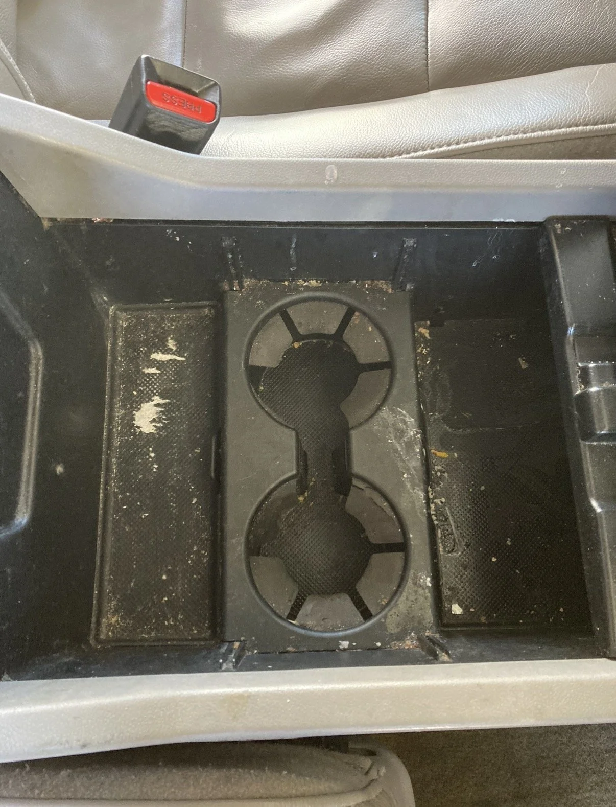 The interior of a vehicle's center console with dusty and dirty cup holders and storage compartments.
