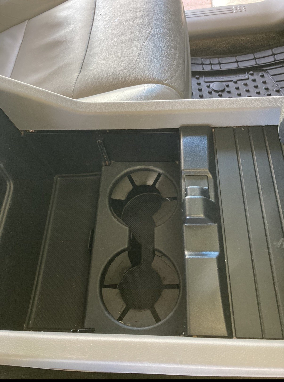 Center console of a vehicle showing cup holders, a gear shift area, and a rubber floor mat. Part of a beige car seat is visible.