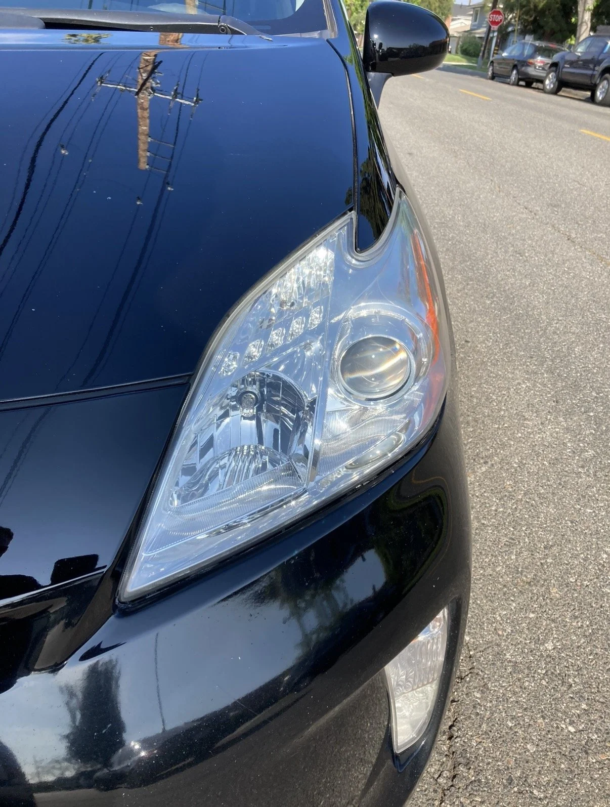 Close-up of the front right side of a black car, showing the headlight and part of the hood with reflections of power lines and a utility pole.