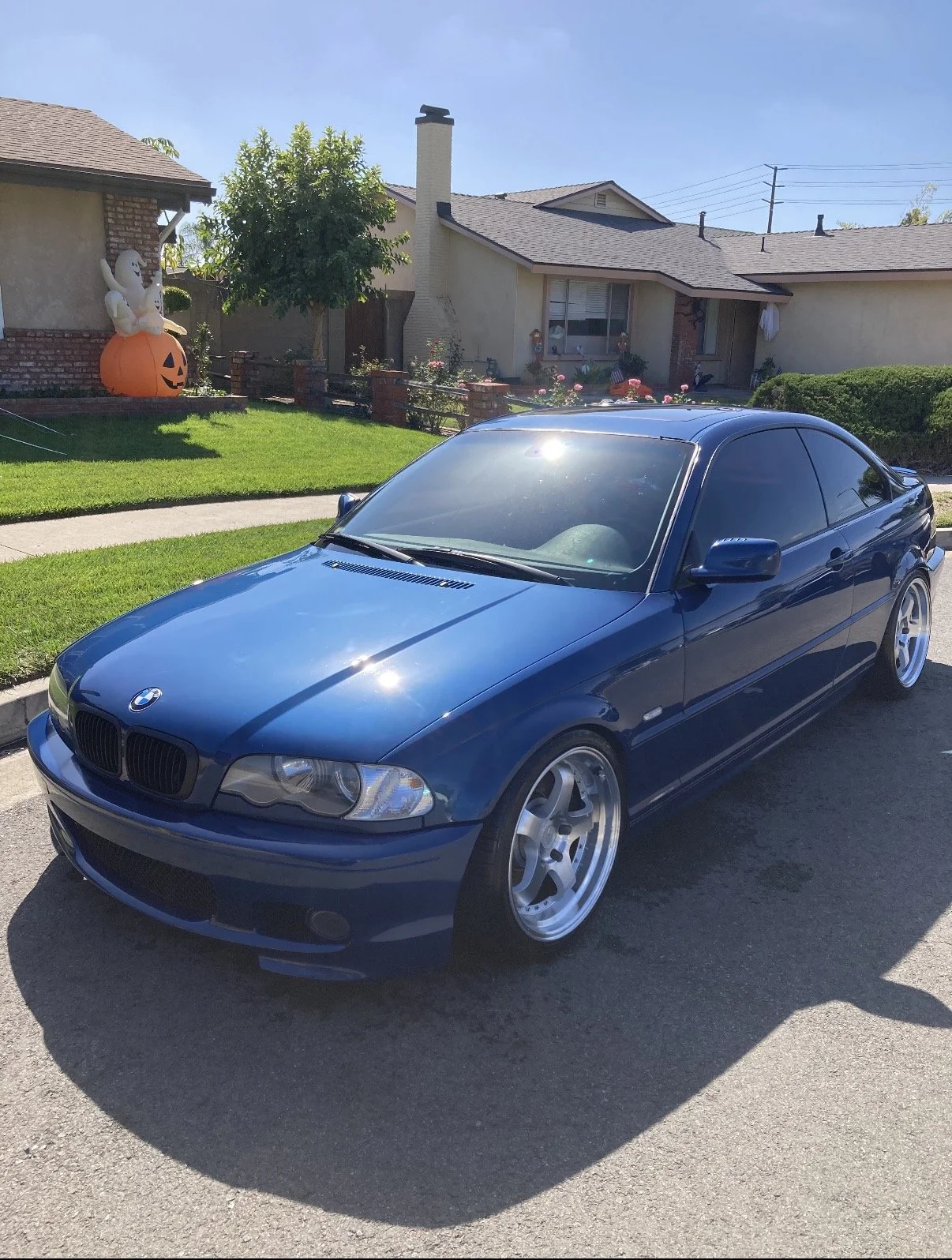 A blue BMW coupe parked on a suburban street with a house and Halloween decorations, including a pumpkin and a ghost figure, in the yard.