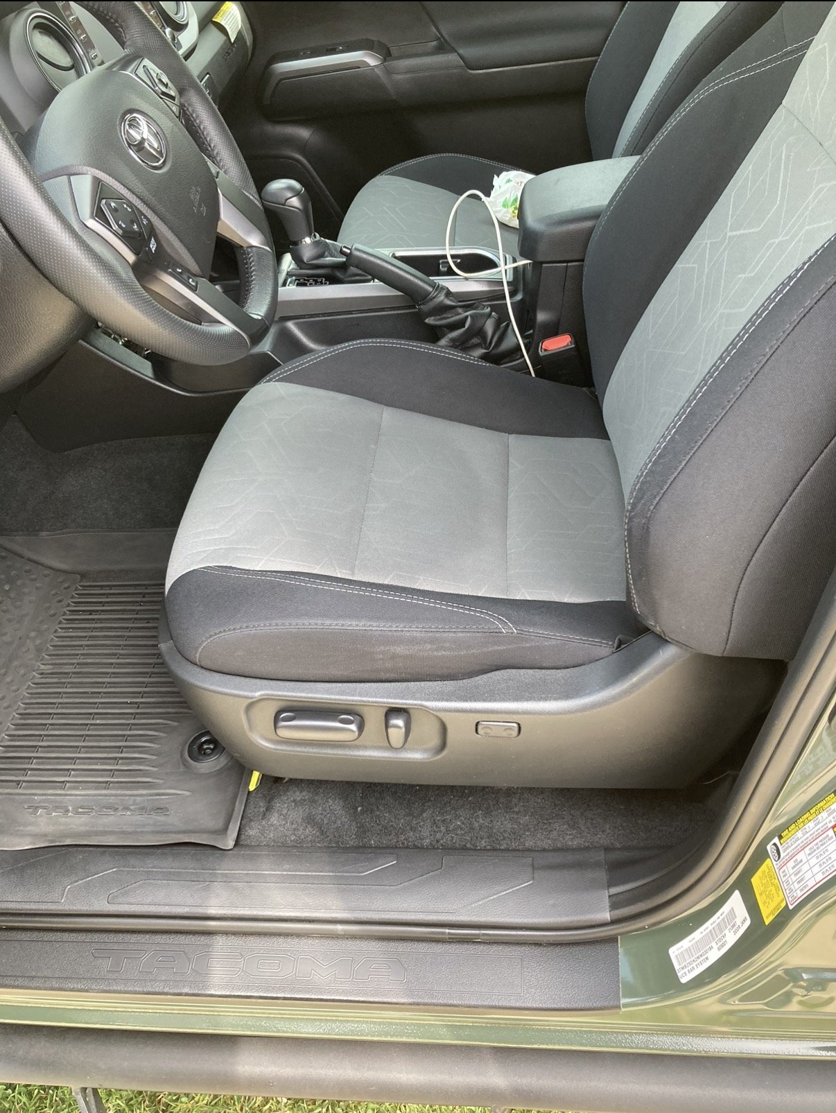 Interior of a Toyota Tacoma truck showing the driver's seat, steering wheel, and part of the front passenger seat. The seats are black and gray with stitching. There is a gear shift and charging cable on the center console.