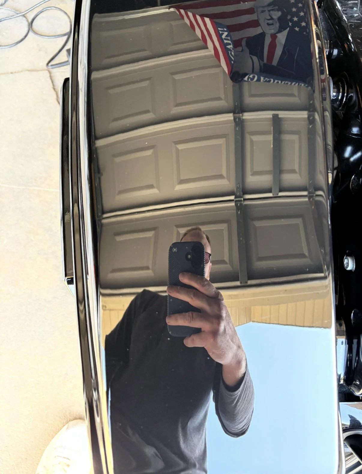 Person taking a selfie in a mirror, holding a phone with a black case. Reflection shows a garage door, a partial wall, and a vintage car with a bumper sticker depicting a political figure.
