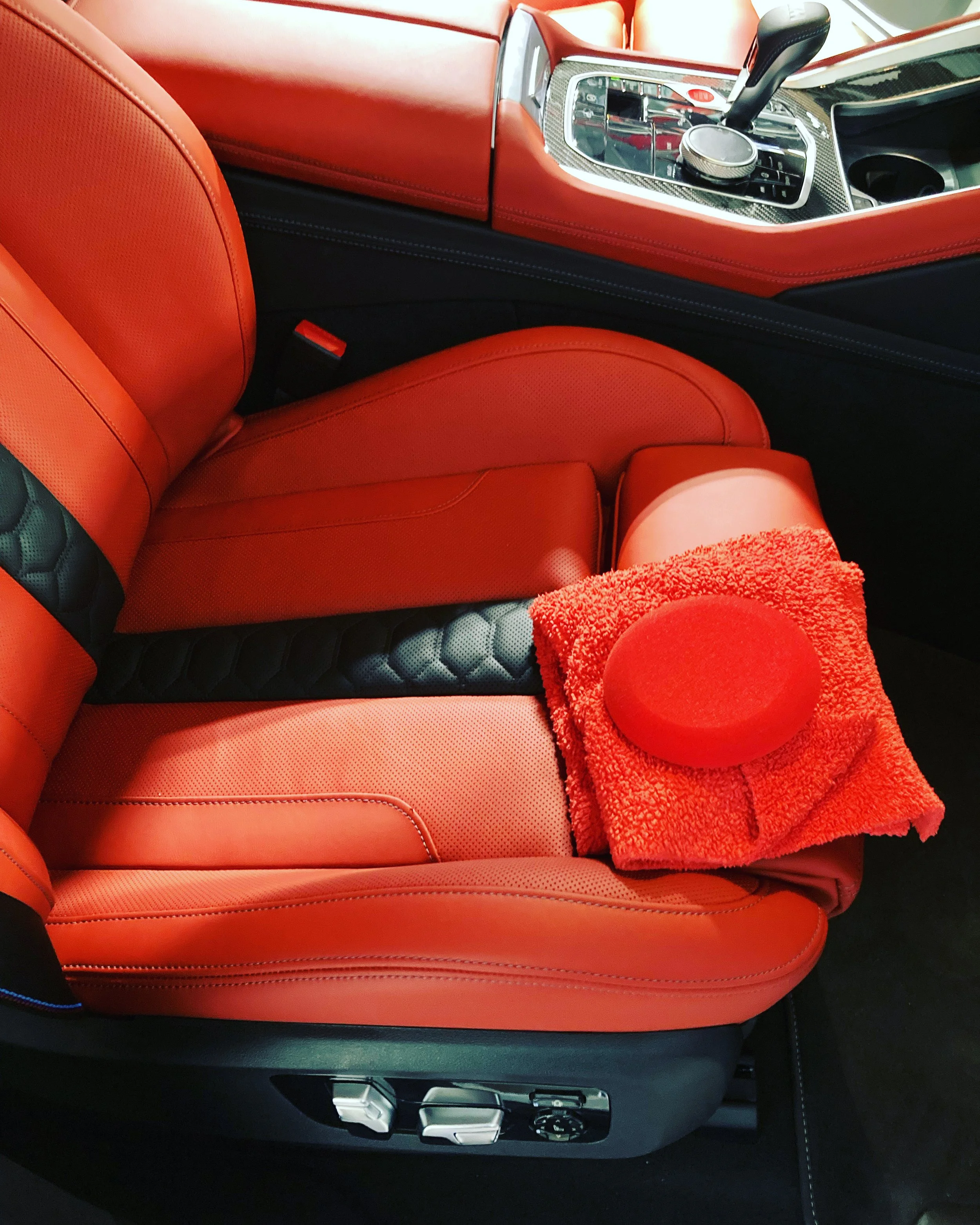 Close-up of a red leather car seat with a matching red towel and a red cap resting on it. The car interior features a center console with various controls and a gear shifter.