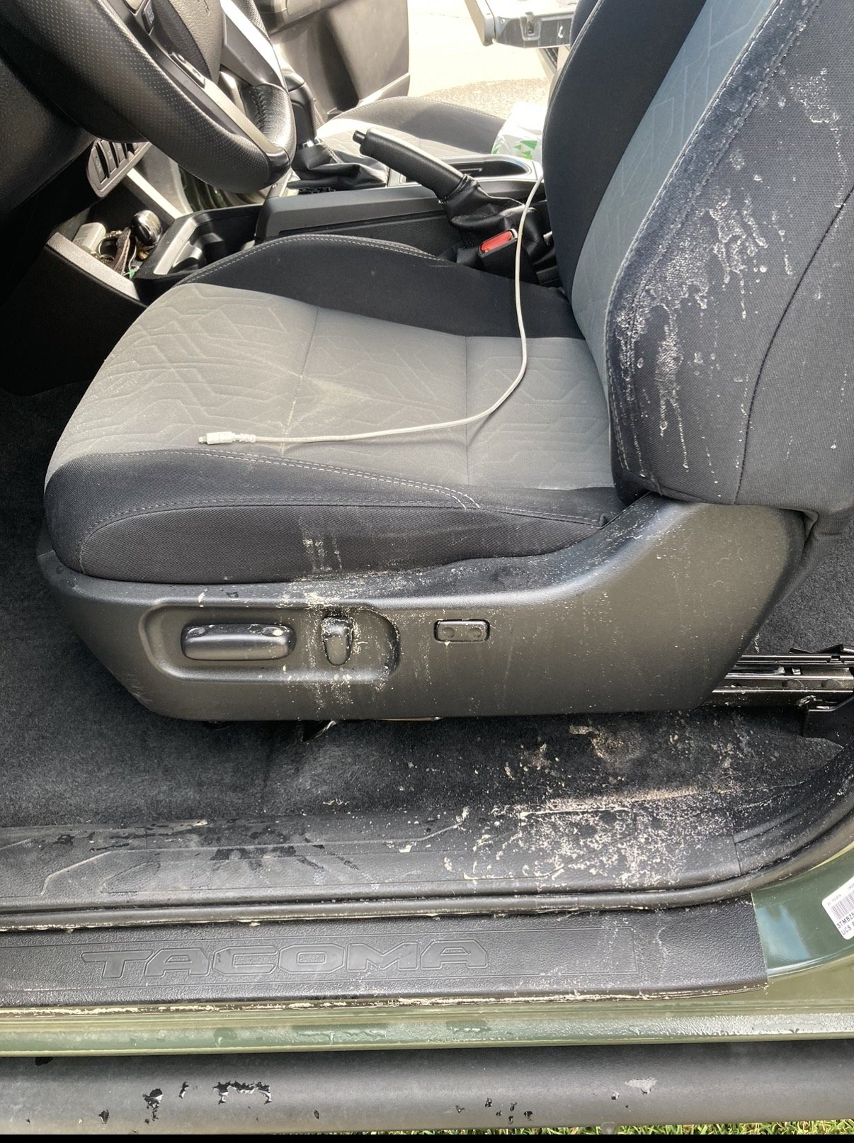 The interior of a Toyota Tacoma truck showing a black fabric driver’s seat with white splatters, a steering wheel, and part of the dashboard. The floor and side panel have white splatters and dirt. A white charging cable is on the seat.