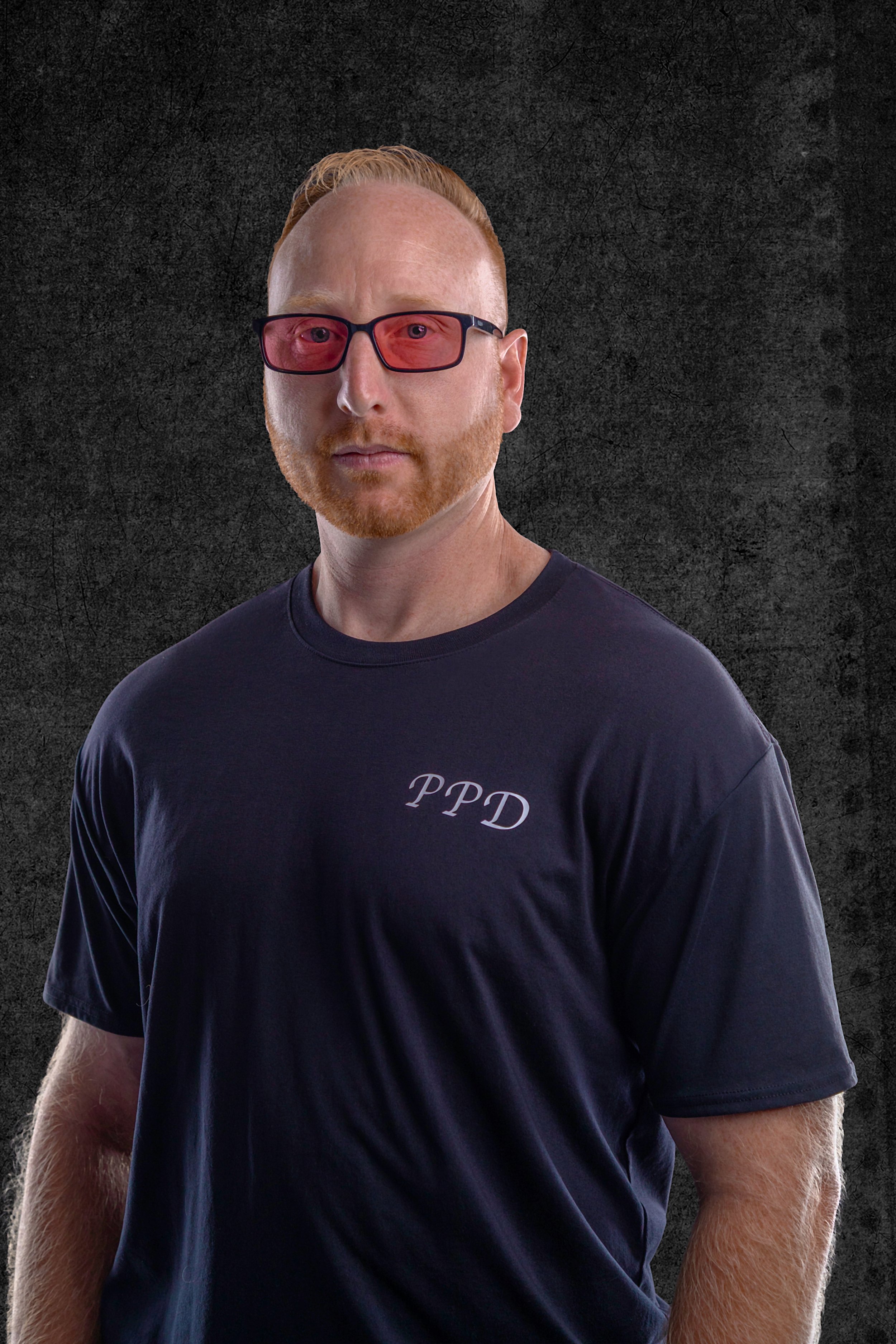 A man with red hair, wearing red-tinted glasses and a navy T-shirt with the initials 'PPD' on it, standing against a dark textured background.