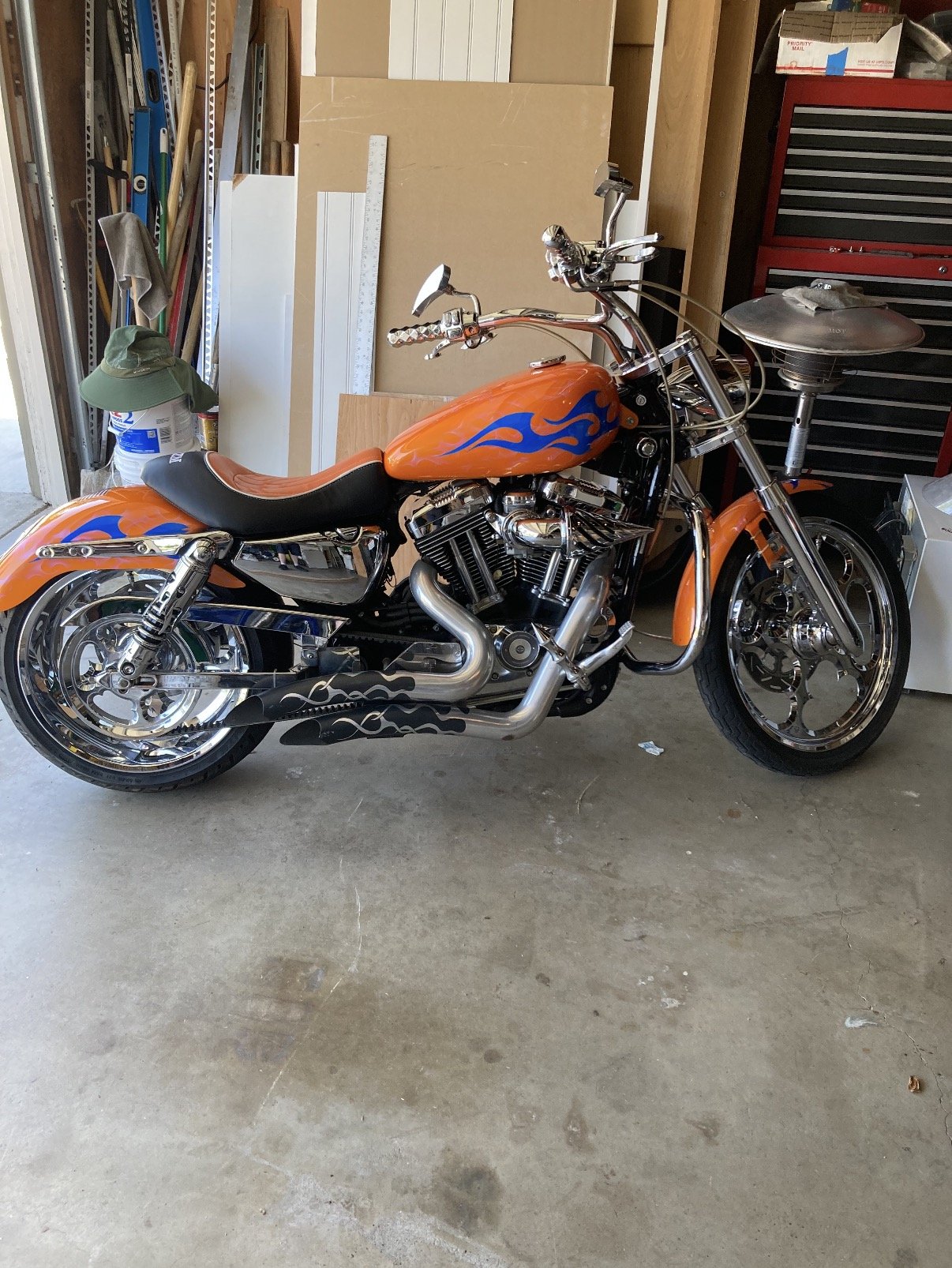 Orange motorcycle with blue flames parked in a garage, surrounded by tools and storage items.