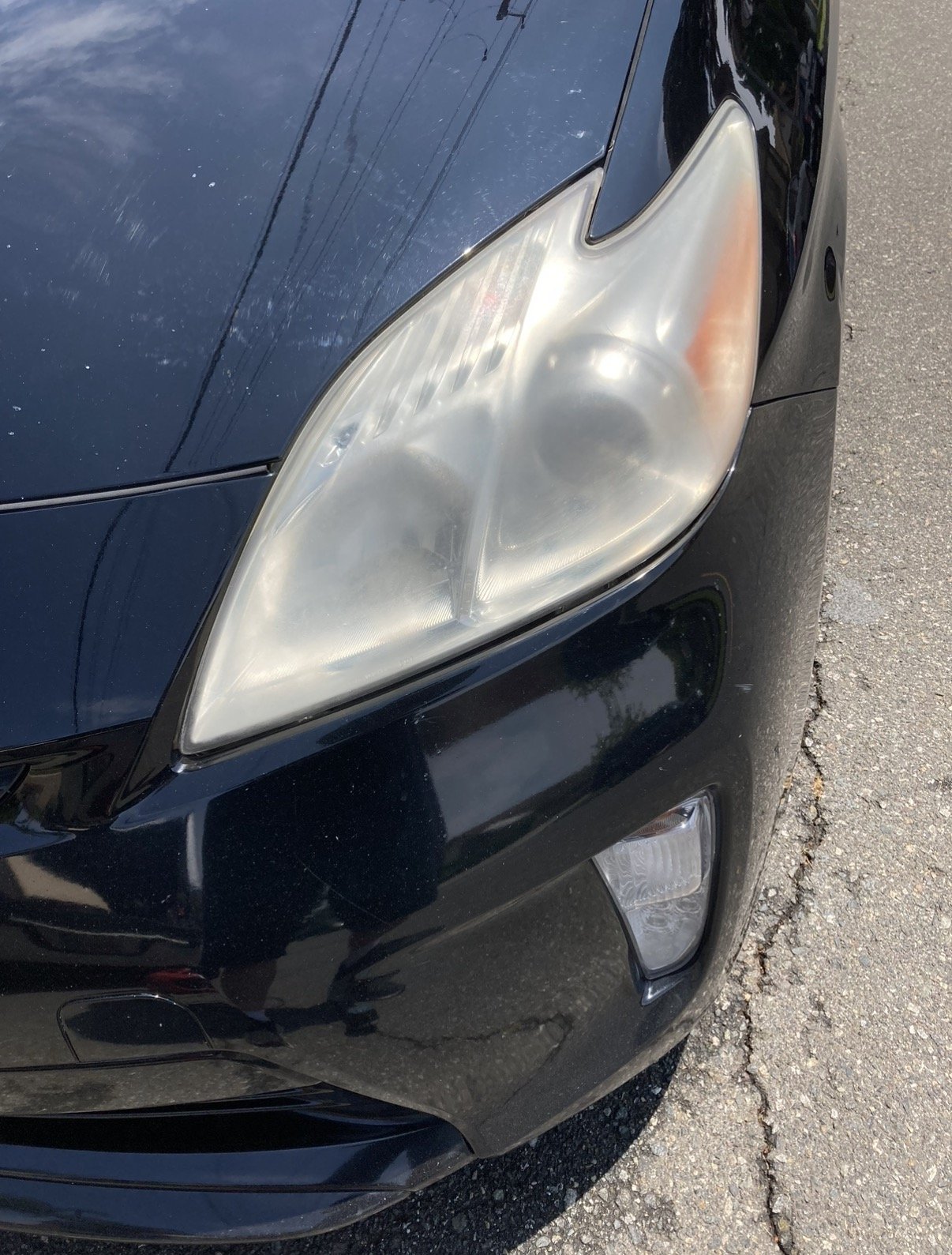 Close-up of a black car's front left headlight and bumper, with some scratches and dirt on the headlight and the road surface visible.