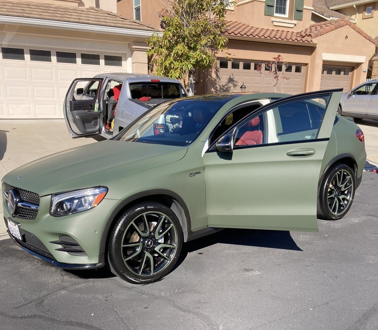 Green Mercedes-Benz sedan with open driver door parked in a residential driveway, with a silver SUV with open side door in the background and houses with garages behind.