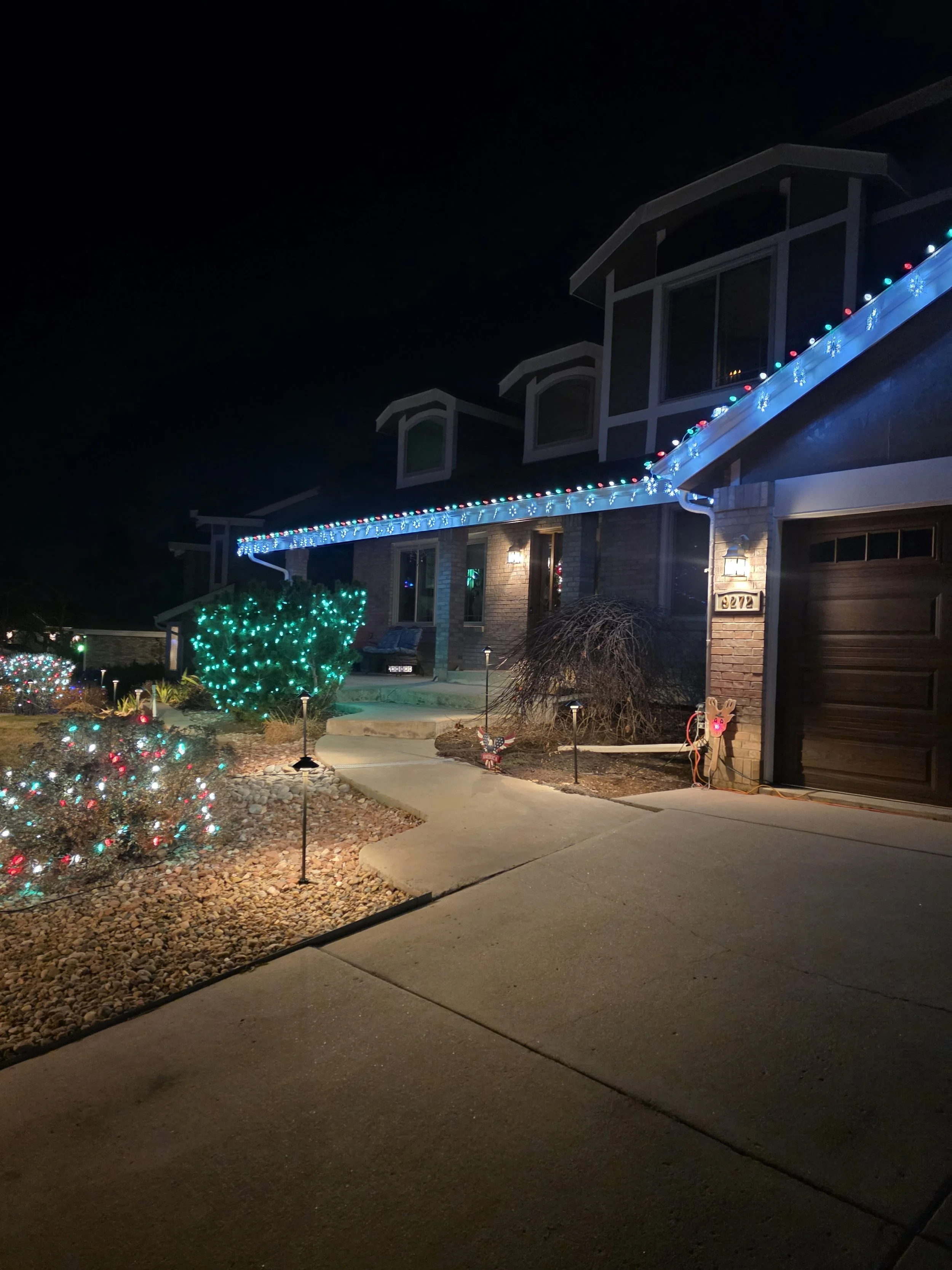 A house with holiday Christmas lights, including blue and red lights on the roof, green lights on bushes, and a lit window. The house has a brick exterior, a garage, and a concrete sidewalk in front.