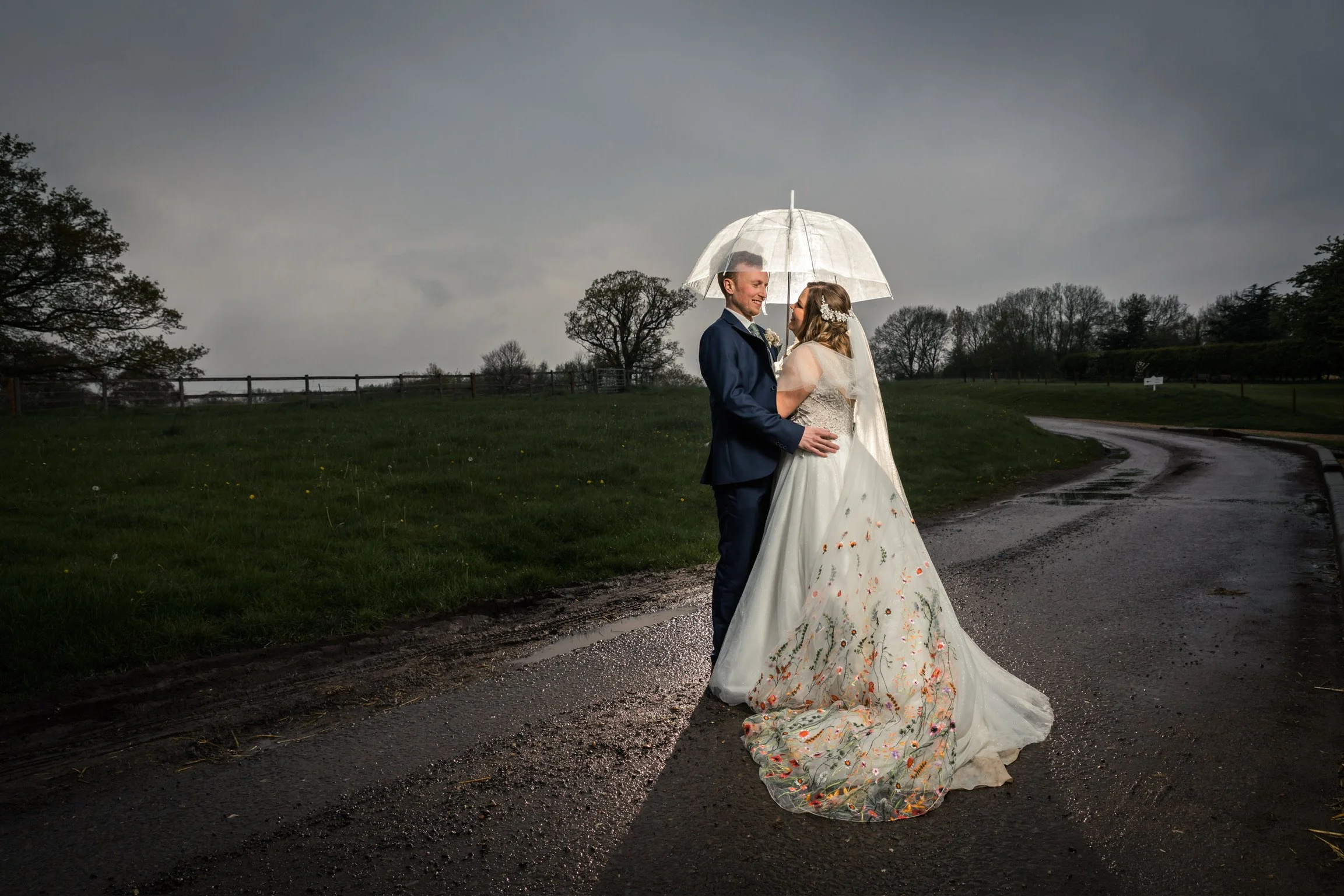 Bride and groom stand looking adoringly at each other under a white umbrella. They are glowing whereas the surroundings are dark and rainy.