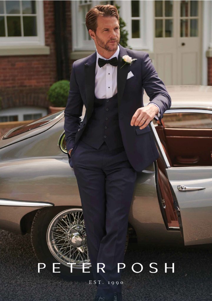 Man in a navy tuxedo with a bow tie and white shirt, standing by a classic car in front of a house, with a white boutonniere and gold watch, looking to the side.